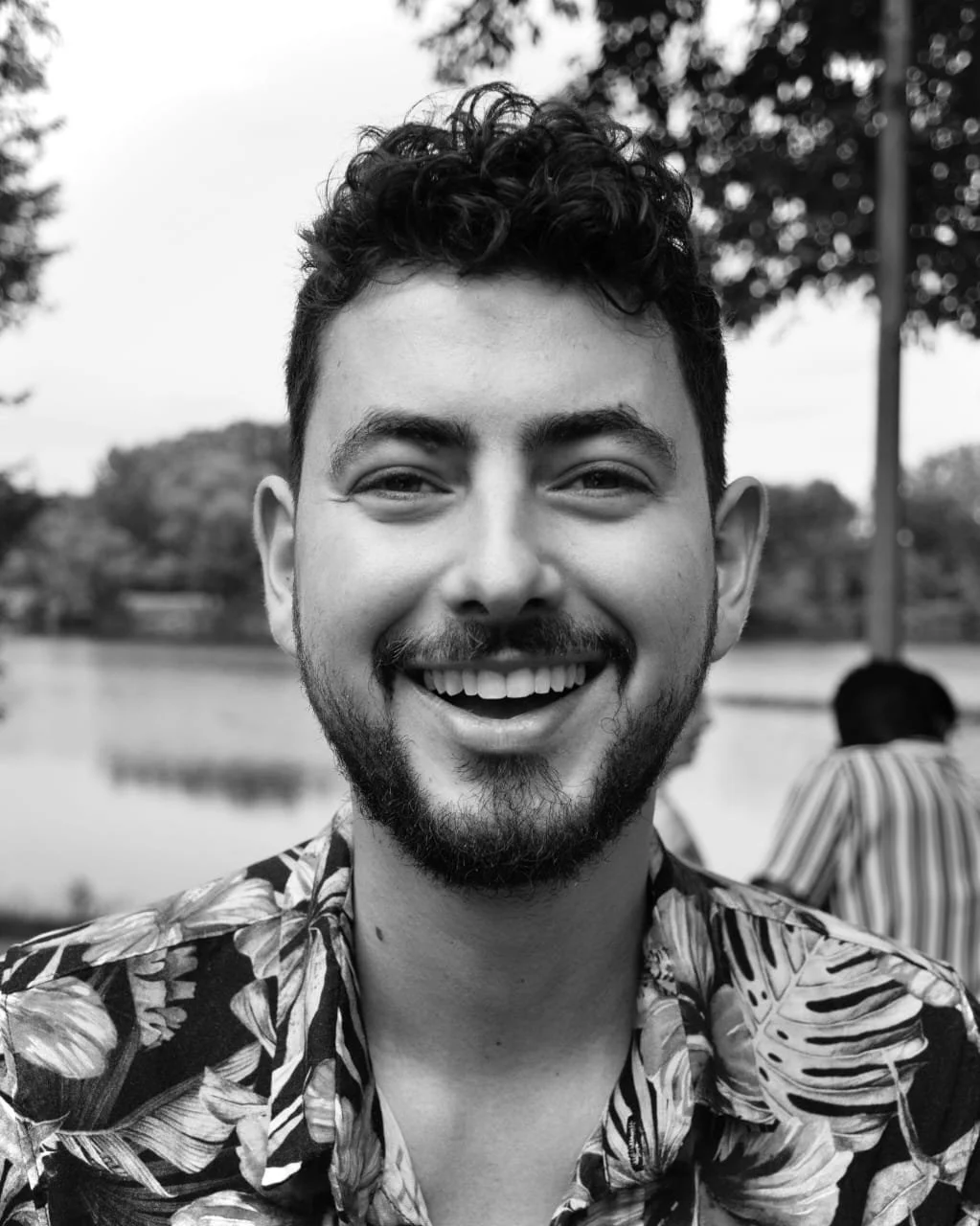 A smiling young man with curly hair and a beard, wearing a floral shirt, outdoors near a body of water with trees in the background.