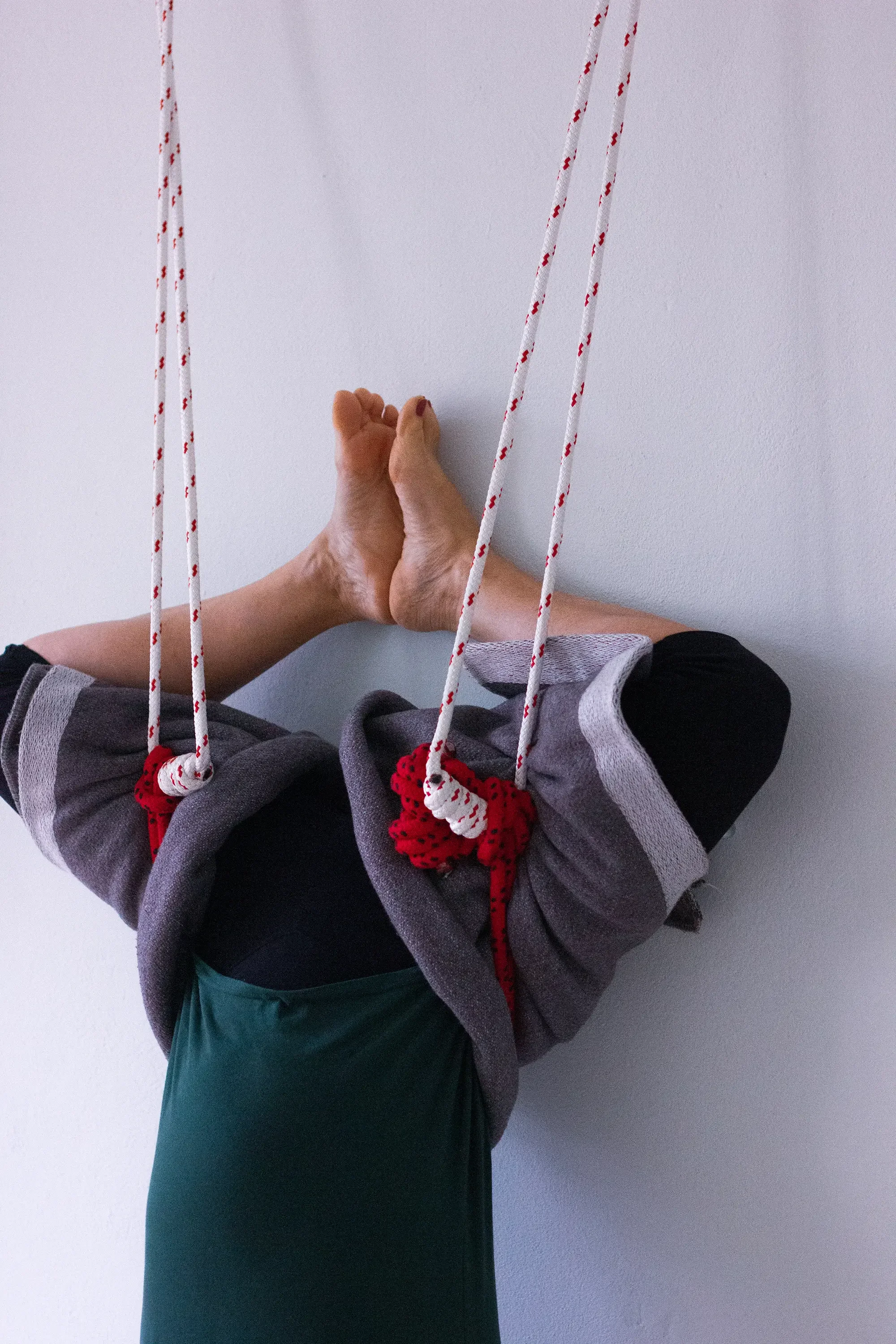Eine Person, die Aerial Yoga praktiziert oder sich dehnt, mit gekreuzten Beinen und gegen die Wand gedrückten Füßen, in einer Schlinge hängend mit roten und weißen Seilen.