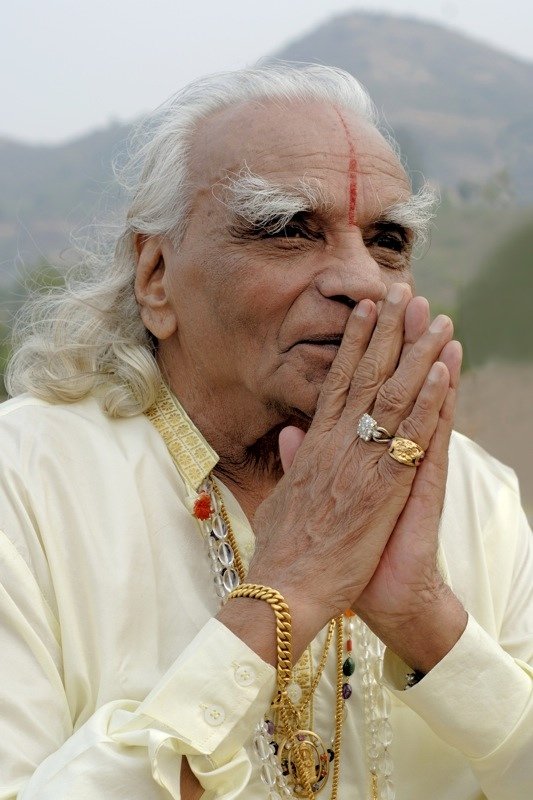 B.K.S. Iyengar trägt traditionelle indische Kleidung und Schmuck, hält seine Hände in einer Gebetshaltung, mit einem roten Zeichen auf der Stirn und einem spirituellen Hintergrund.