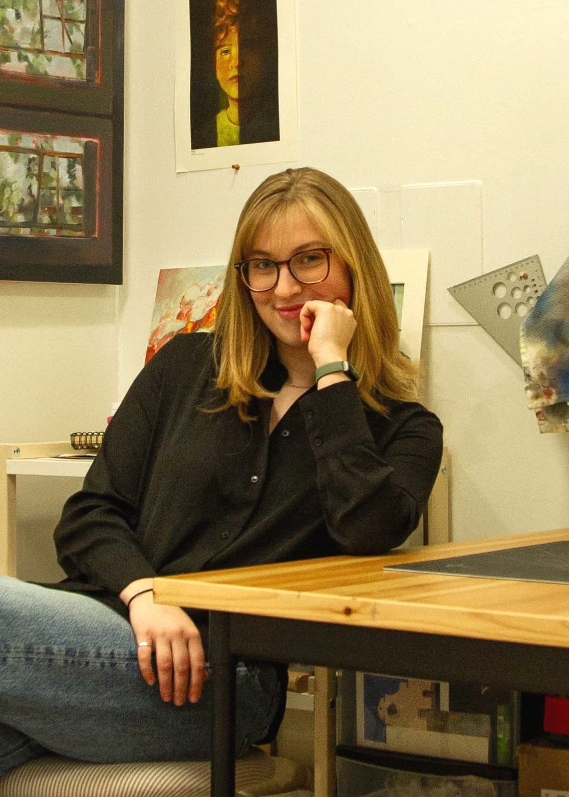 A woman with blonde hair, glasses, and a black shirt sitting at a desk in an art studio, resting her chin on her hand with a slight smile.