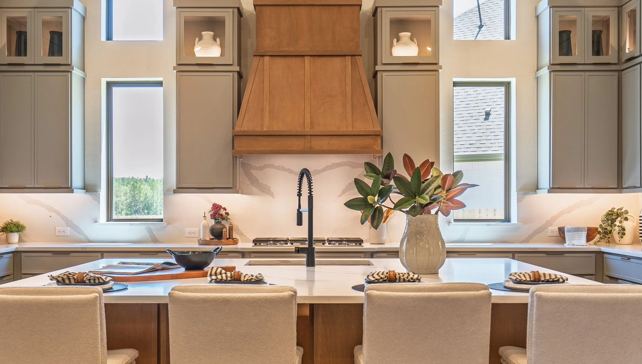 Modern kitchen with beige cabinetry, a white countertop island, a large brown range hood, three windows behind the counter, potted plants, and a vase with large green leaves in the foreground.