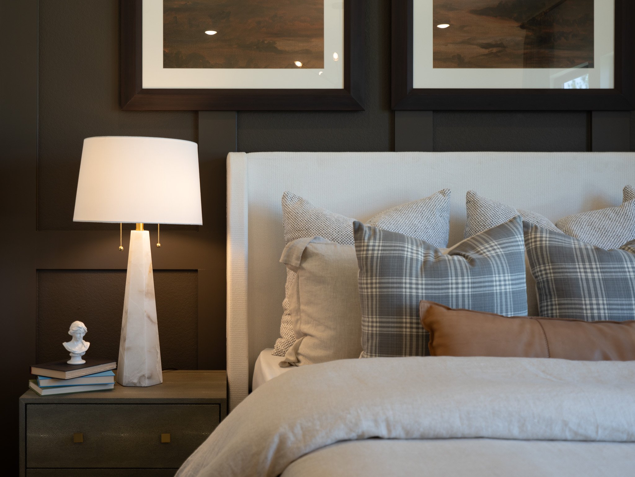 A bedroom with a white upholstered bed, checkered pillows, a dark nightstand with a small white bust sculpture, a lamp with a white shade, two framed pictures on the dark wall, and a beige blanket at the foot of the bed.