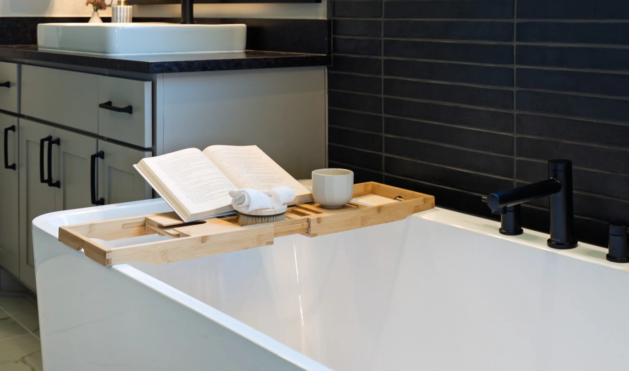 A modern bathroom featuring a white bathtub with a wooden bath tray holding an open book, a white mug, a small brush, and a rolled towel. The background shows a vanity with a vessel sink, a black faucet, and dark tiled walls.