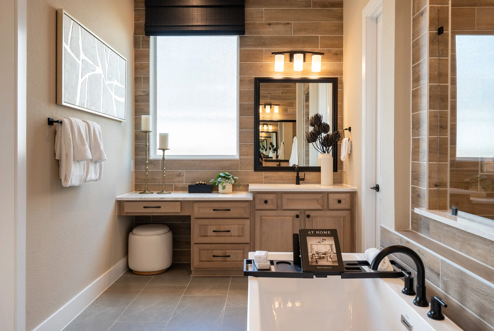 A modern bathroom with beige wooden cabinets, a black framed mirror, a black faucet, a small plant, and decorative candles near a frosted window. The walls are tiled with horizontal wood-look tiles, and there are white towels on a black towel bar.