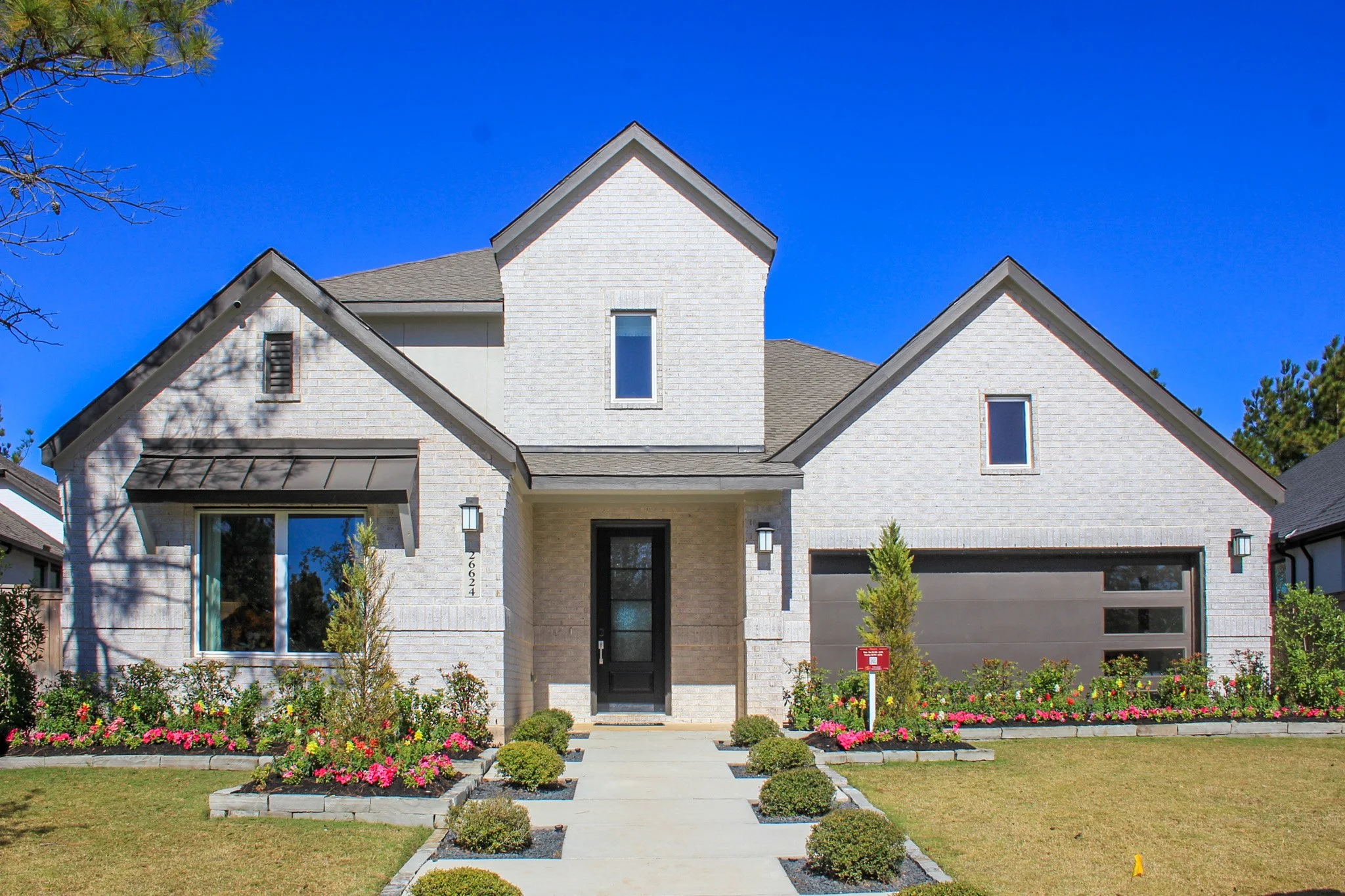 Modern white brick house with a gabled roof, front lawn with flower bed, small trees, and a paved walkway leading to the front entrance, under a bright blue sky.