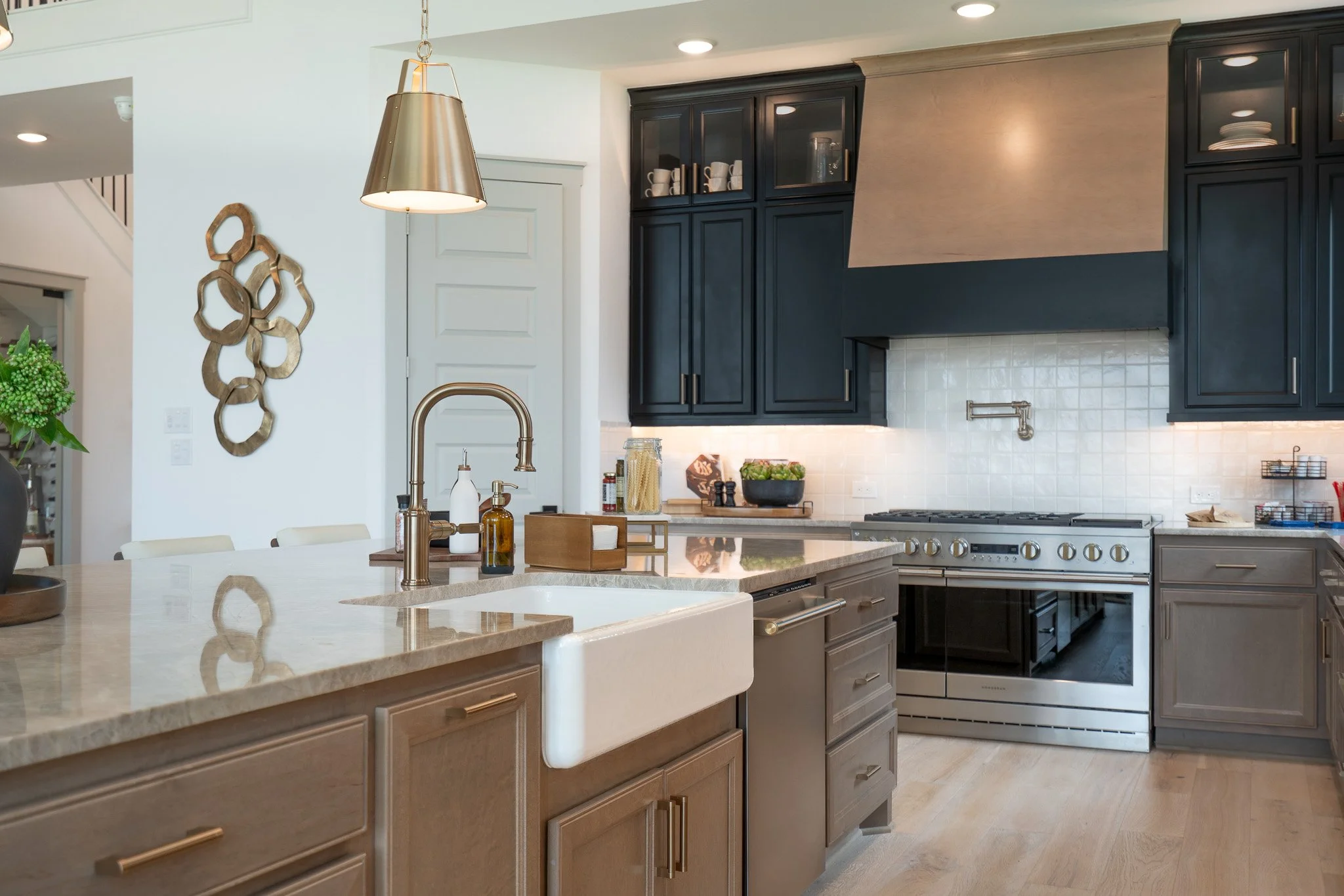 Modern kitchen with island, beige countertop, black upper cabinets, white tiled backsplash, and stainless steel stove; pendant light and decorative wall art.