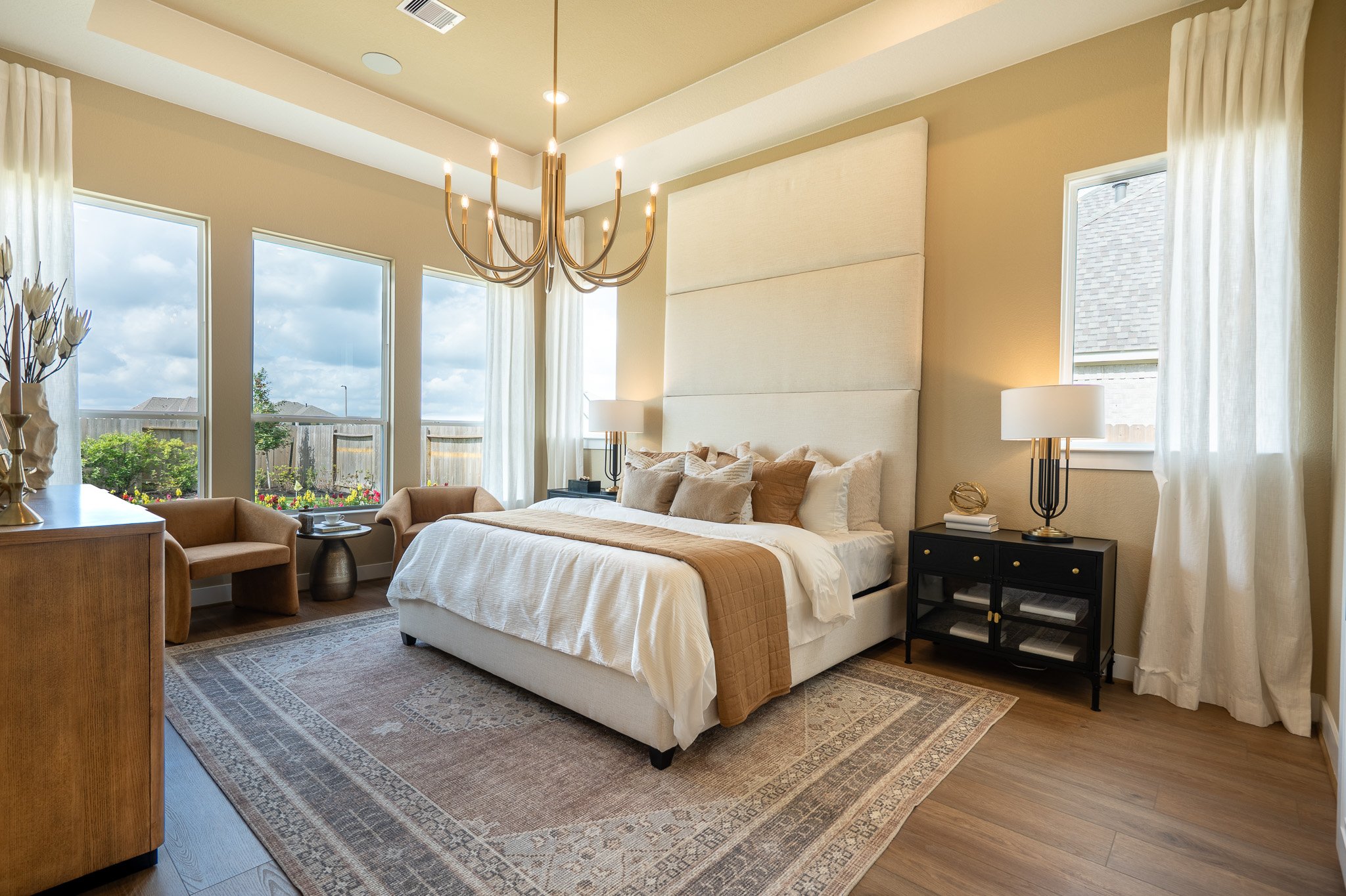A bedroom featuring three large windows, a bed with pillows and a beige cover, a black nightstand with a lamp, two beige armchairs, a stainless steel vase, and a chandelier hanging from the ceiling.
