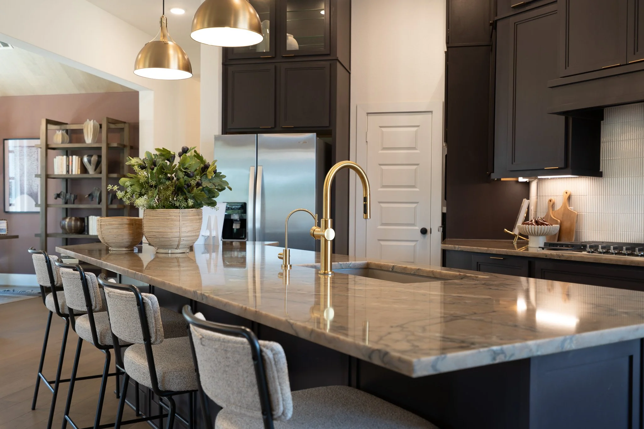 Modern kitchen with black cabinets, a marble countertop island, gold fixtures, plants, and a bookshelf in the background.