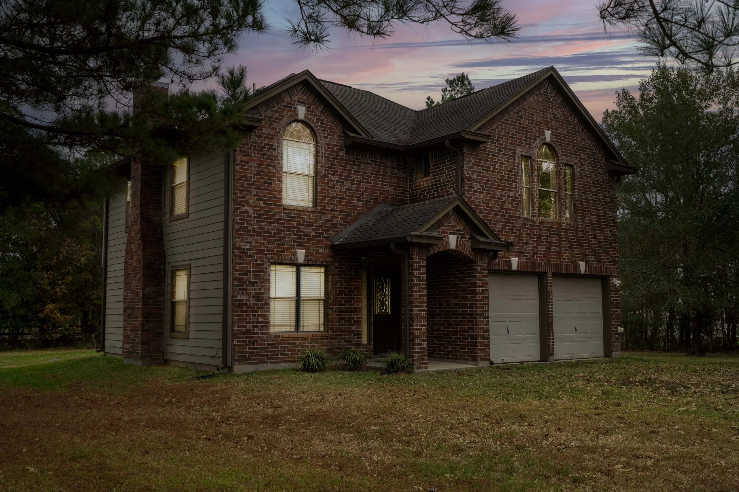 Brick two-story house with attached garage, surrounded by trees, at sunset with colorful sky.
