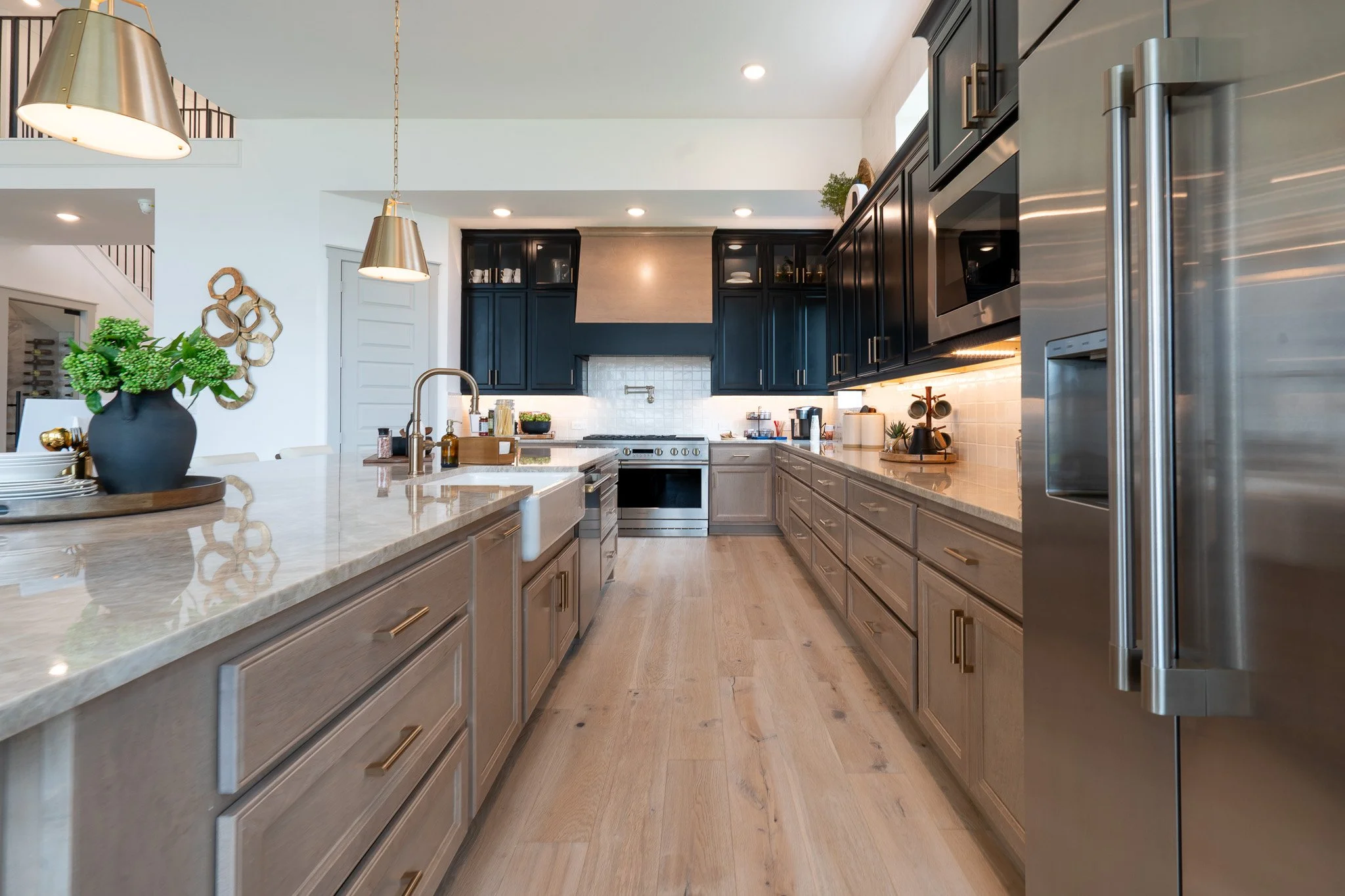 A modern kitchen with beige and dark blue cabinets, stainless steel appliances, a marble island counter with a small plant, gold pendant lights, and wooden flooring.