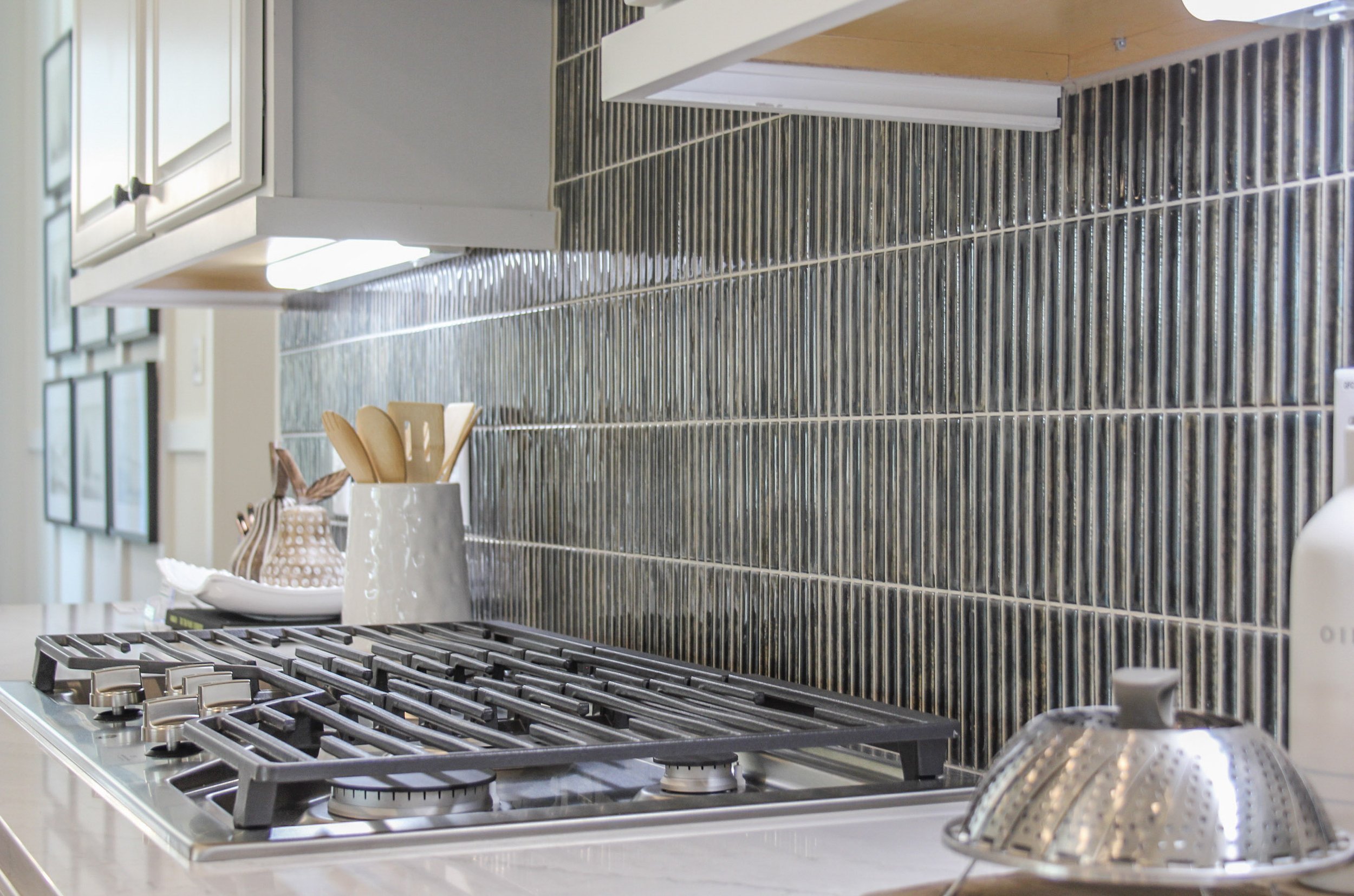 Kitchen with stainless steel stove and black tile backsplash, countertop with a white vase holding wooden utensils, and a wire colander on the counter.