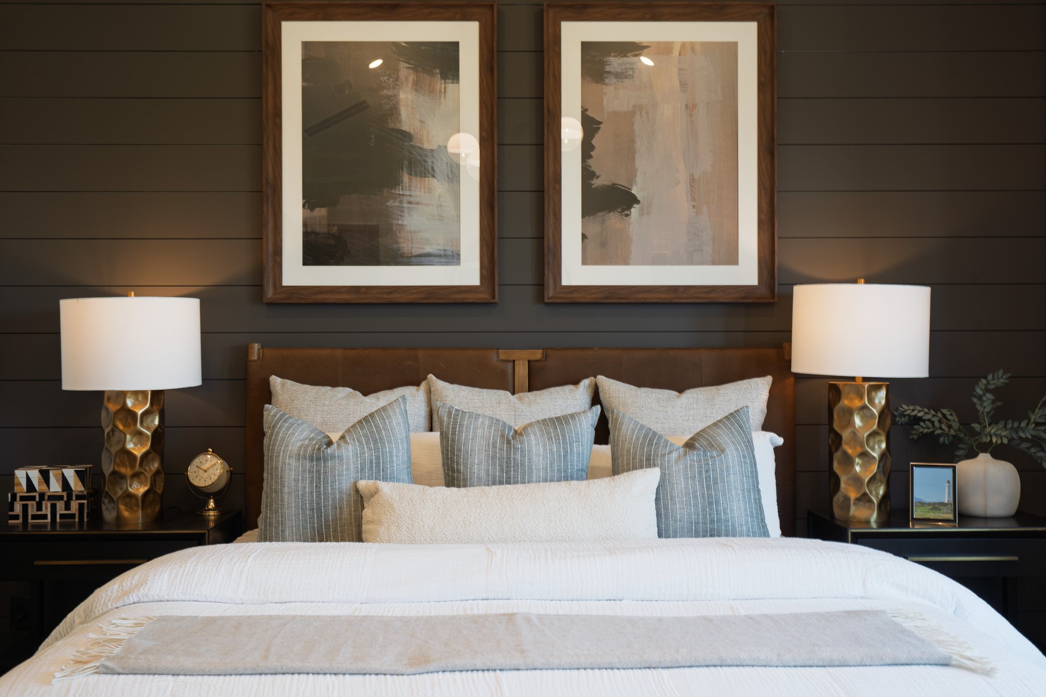 A cozy bedroom with a large bed, gray-striped pillows, and beige bedding. Two matching gold lamps with white shades sit on black nightstands on each side of the bed. Above the bed are two abstract paintings framed in wood, hanging against a dark gray