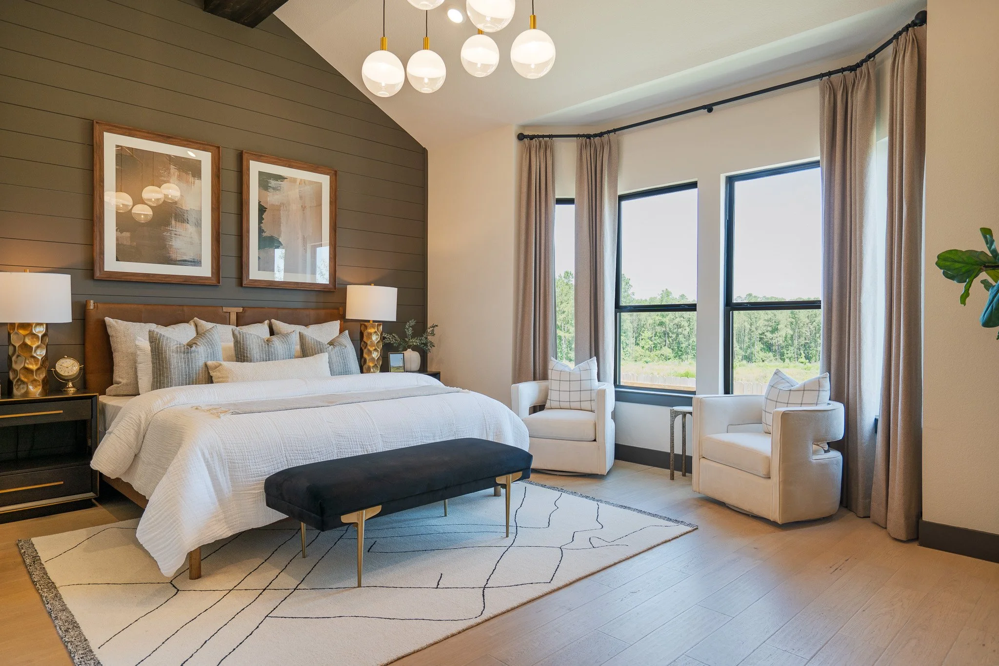 A modern bedroom with a large bed with white bedding, two side tables with lamps, framed artwork above the bed, and large windows with beige curtains. There are two white armchairs and a small side table near the windows, with a view of trees outside