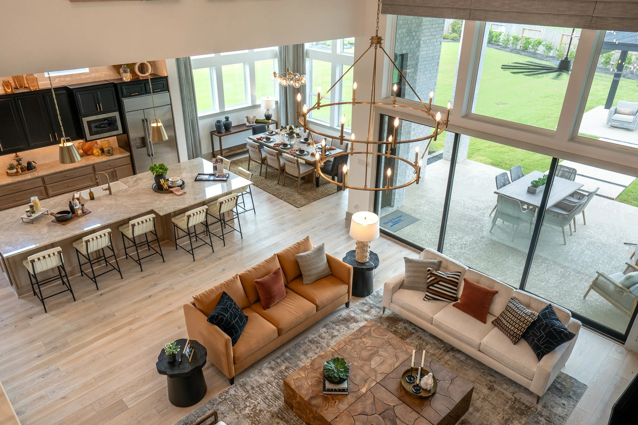 A spacious open-concept living and dining area with large windows overlooking a backyard. The living room has a tan and cream sofa with decorative pillows, a black side table with plants, and a patterned rug. The dining area features a rectangular ta
