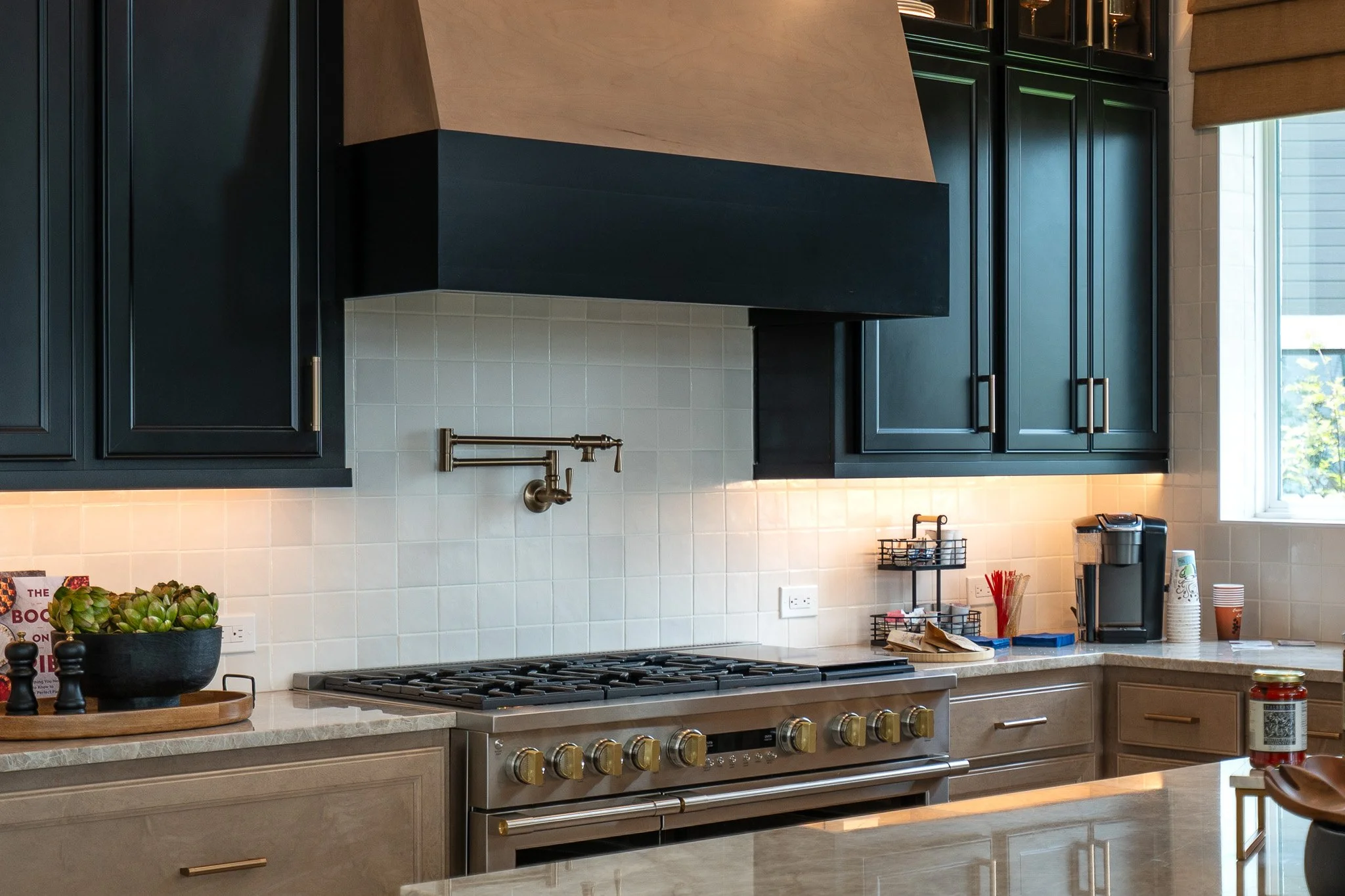 Modern kitchen with black upper cabinets, a stainless steel stove, a beige countertop, a white tiled backsplash, and a window with a view outside.