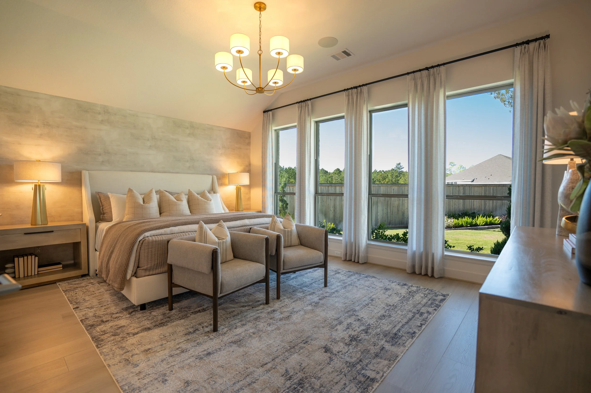Bright living room with large windows, white curtains, a chandelier, a cream-colored sofa, side tables with lamps, and decorative pillows.