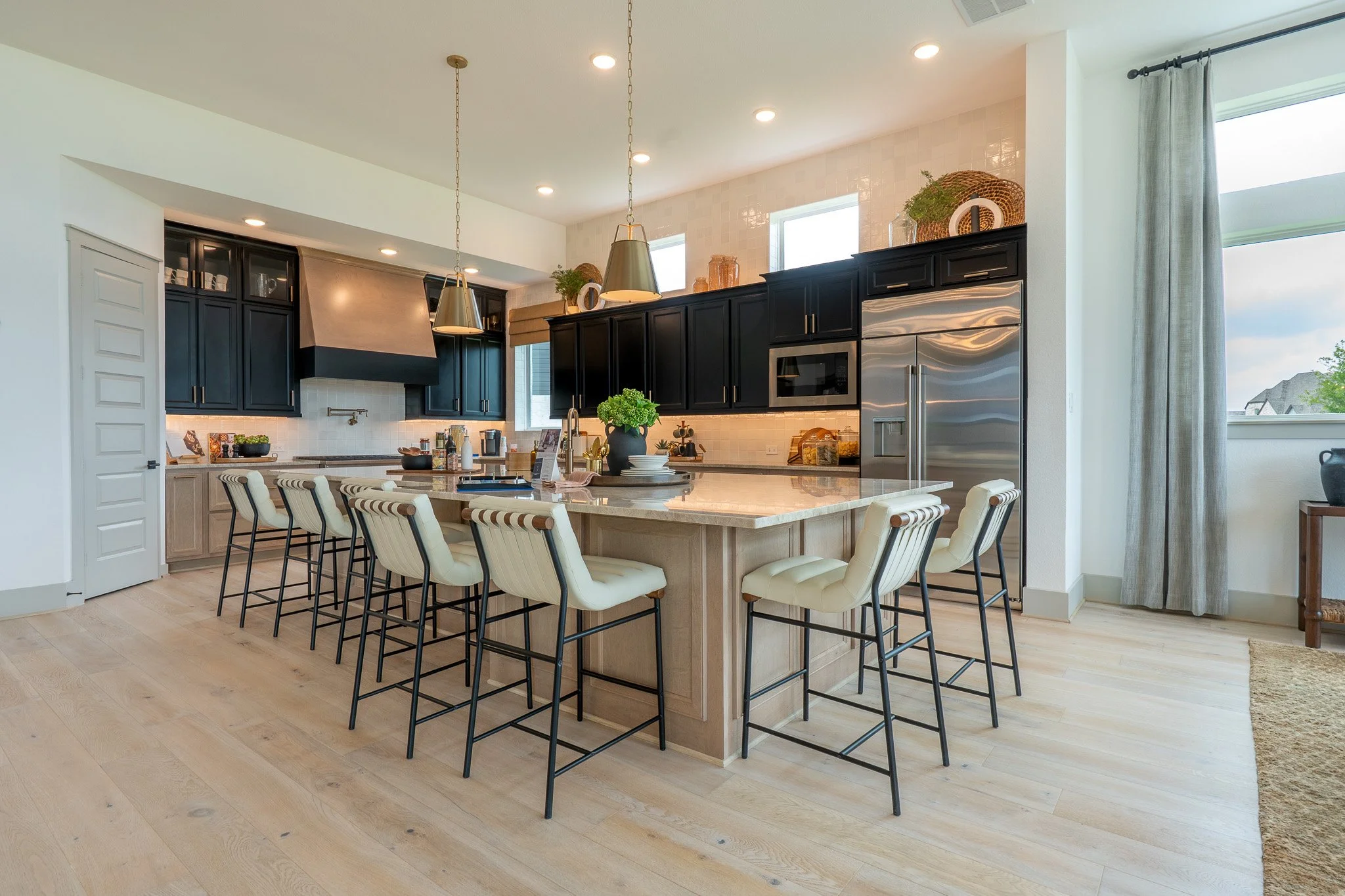 Modern kitchen with black cabinets, large island, white chairs, stainless steel appliances, pendant lights, and hardwood floors.