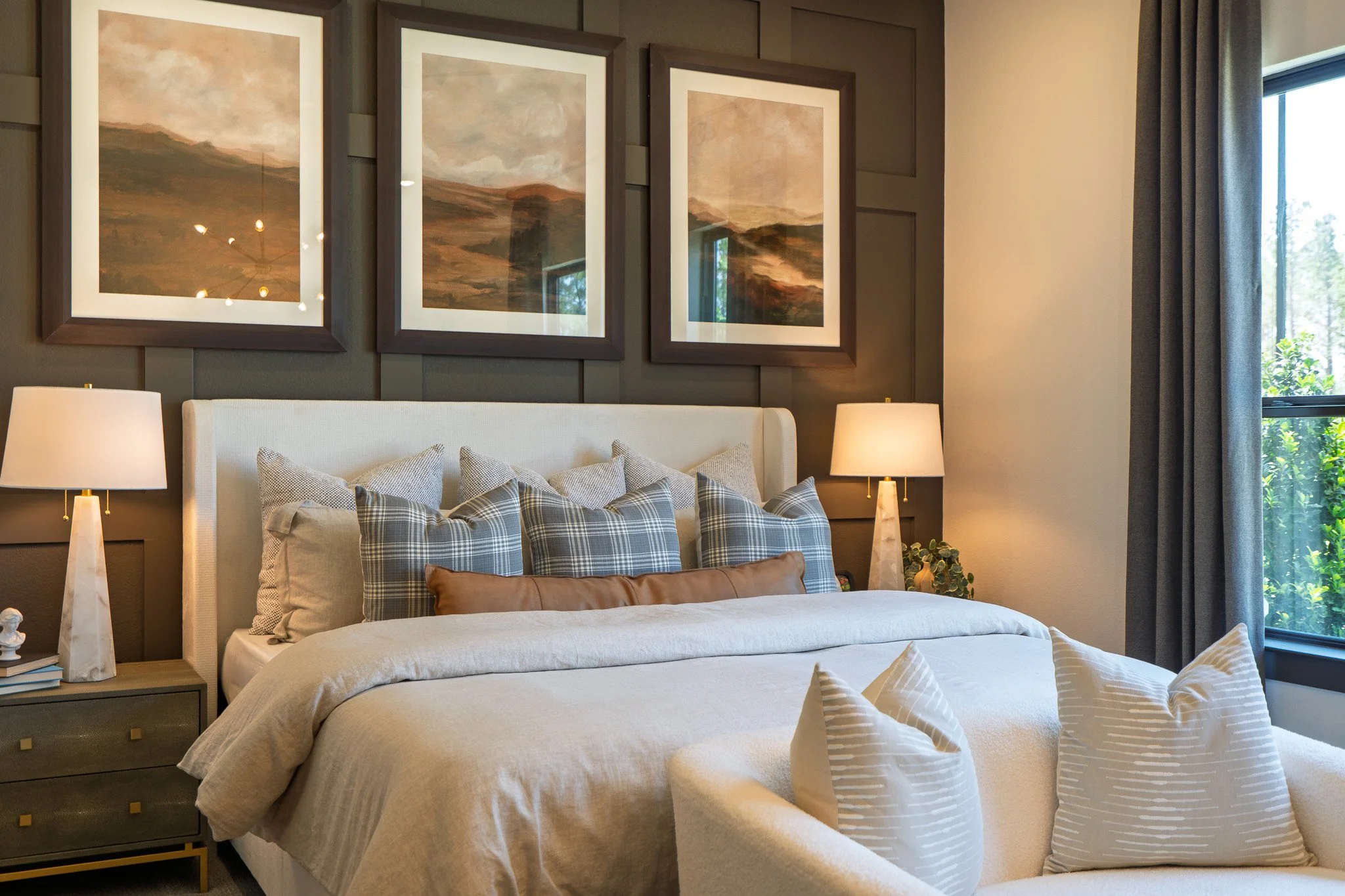 A cozy bedroom with a white bed, multiple patterned pillows, two bedside lamps on dark wood nightstands, three landscape paintings above the bed, and a window with gray curtains showing greenery outside.
