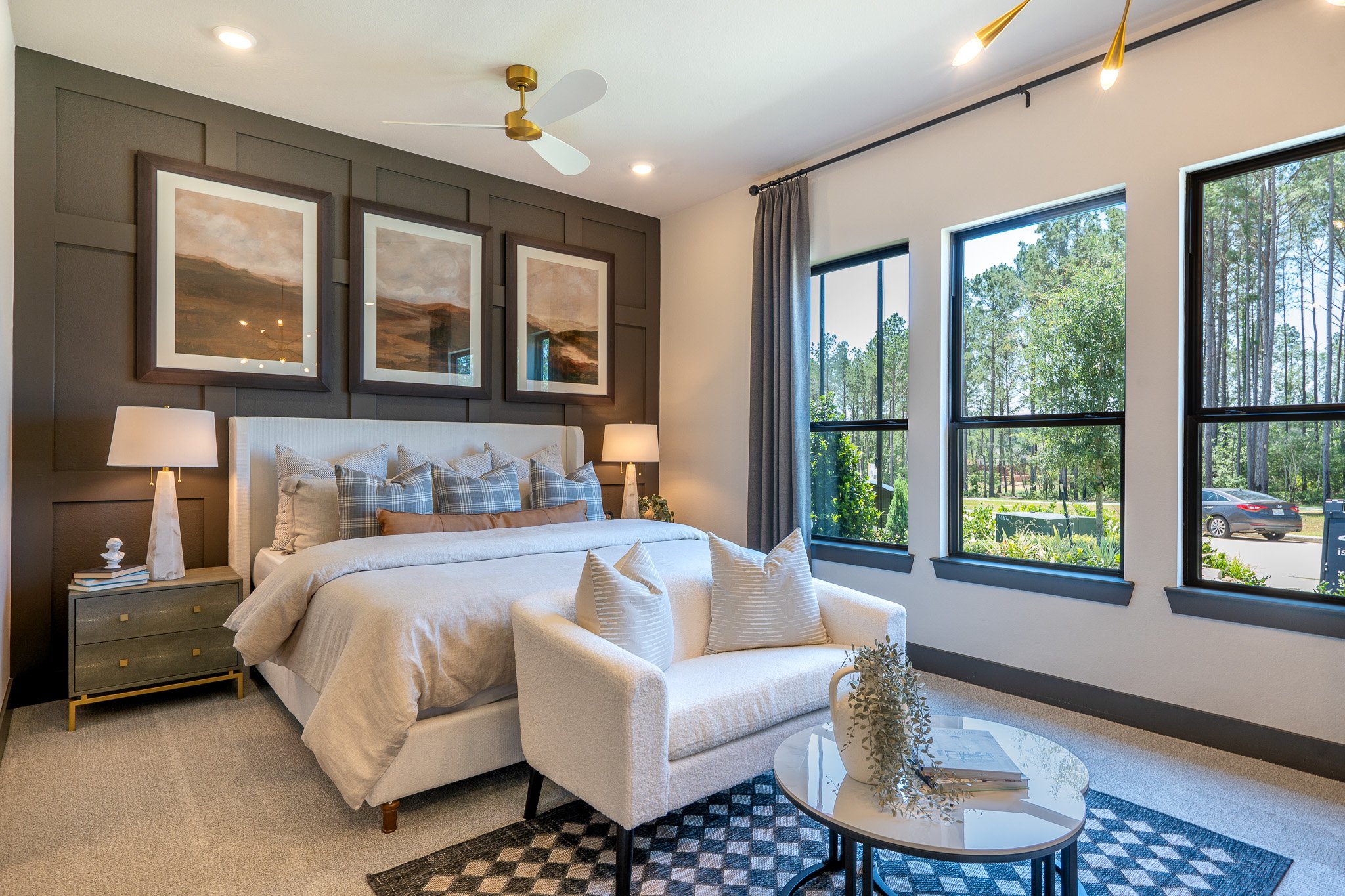 A bedroom with large windows showing a view of trees outside. The room has a white bed with plaid pillows, two lamps on the nightstands, and modern wall art above the bed. There is a white armchair, a round glass table, and a patterned rug in the are