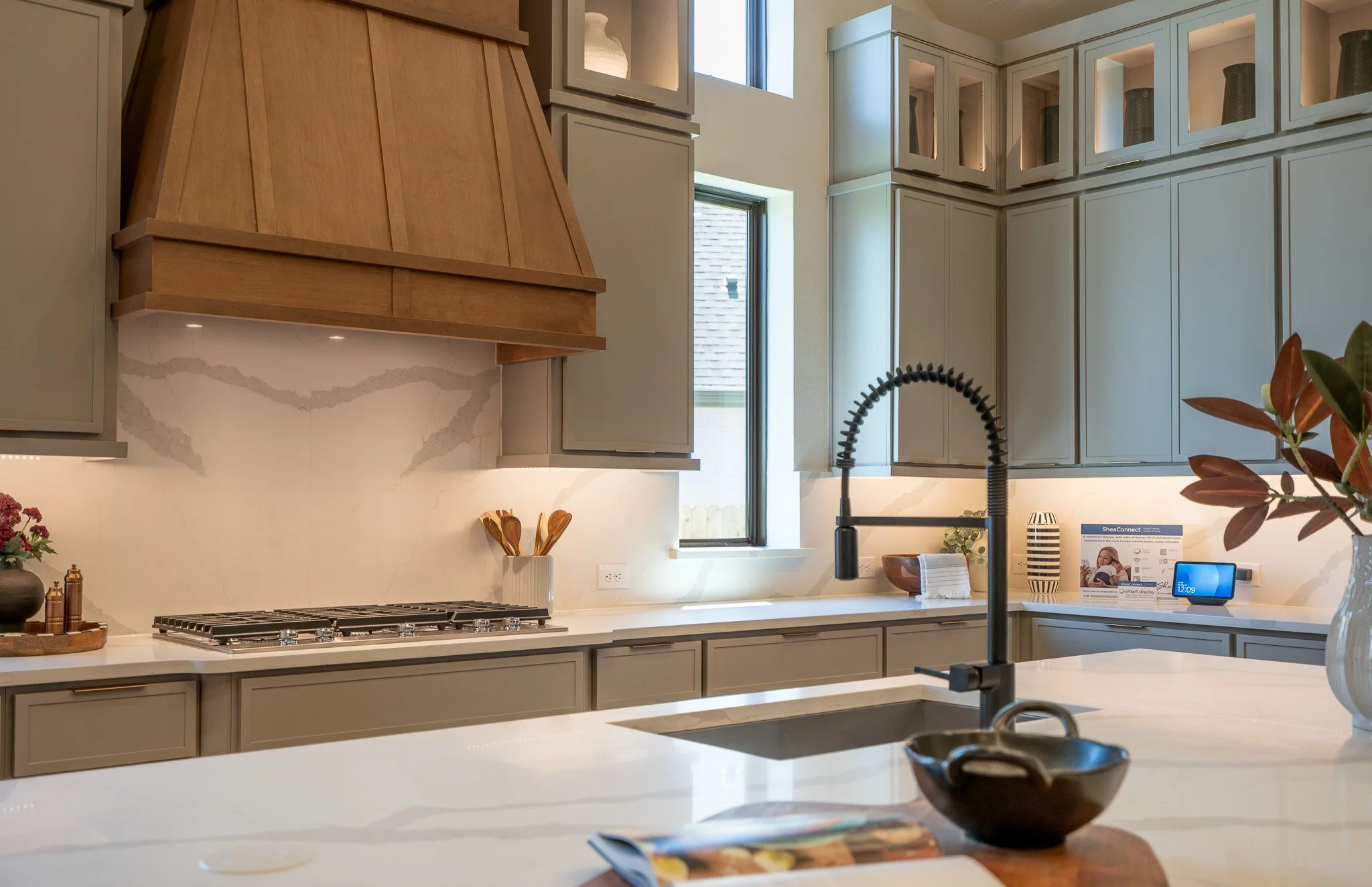Modern kitchen with beige cabinetry, marble backsplash, and black faucet. Includes plant, decorative vases, and digital device on the countertop.