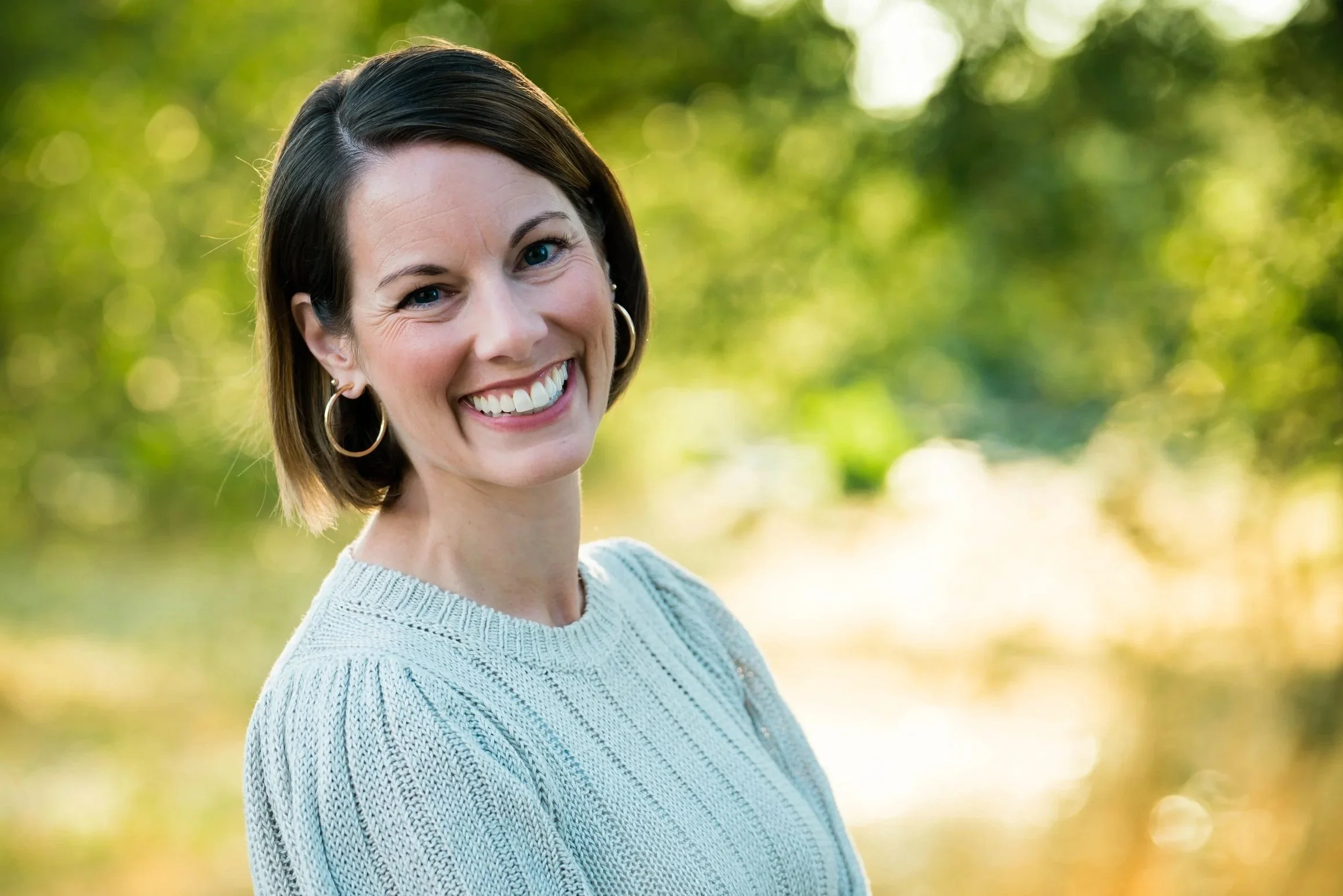 A woman with short brown hair, wearing hoop earrings and a light knit sweater, smiling outdoors with green trees and sunlight in the background.