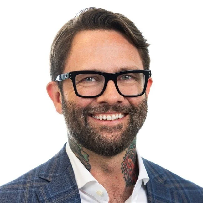 Smiling man with glasses, beard, and a neck tattoo, wearing a suit jacket and white shirt, against a white background.