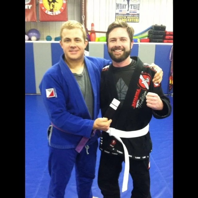 Two men in Brazilian Jiu-Jitsu uniforms, smiling, one holding a black belt, in a gym with mats and training equipment.
