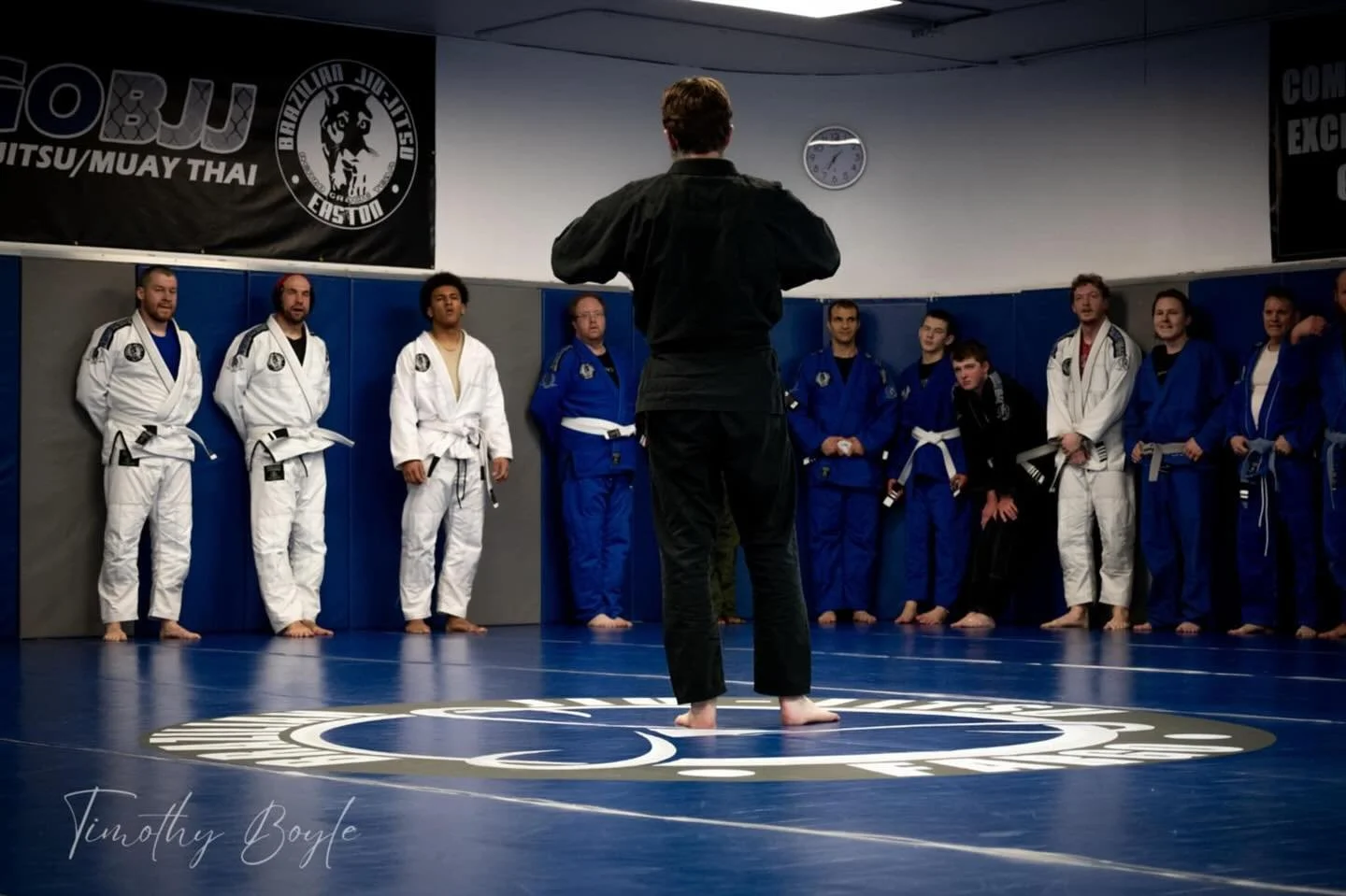 A martial arts class at a gym with students in white and blue gi standing in a semi-circle around an instructor in black gi, who is demonstrating or speaking, on a blue mat with a circular logo. There are banners on the wall, a clock, and a subtle signature at the bottom left.