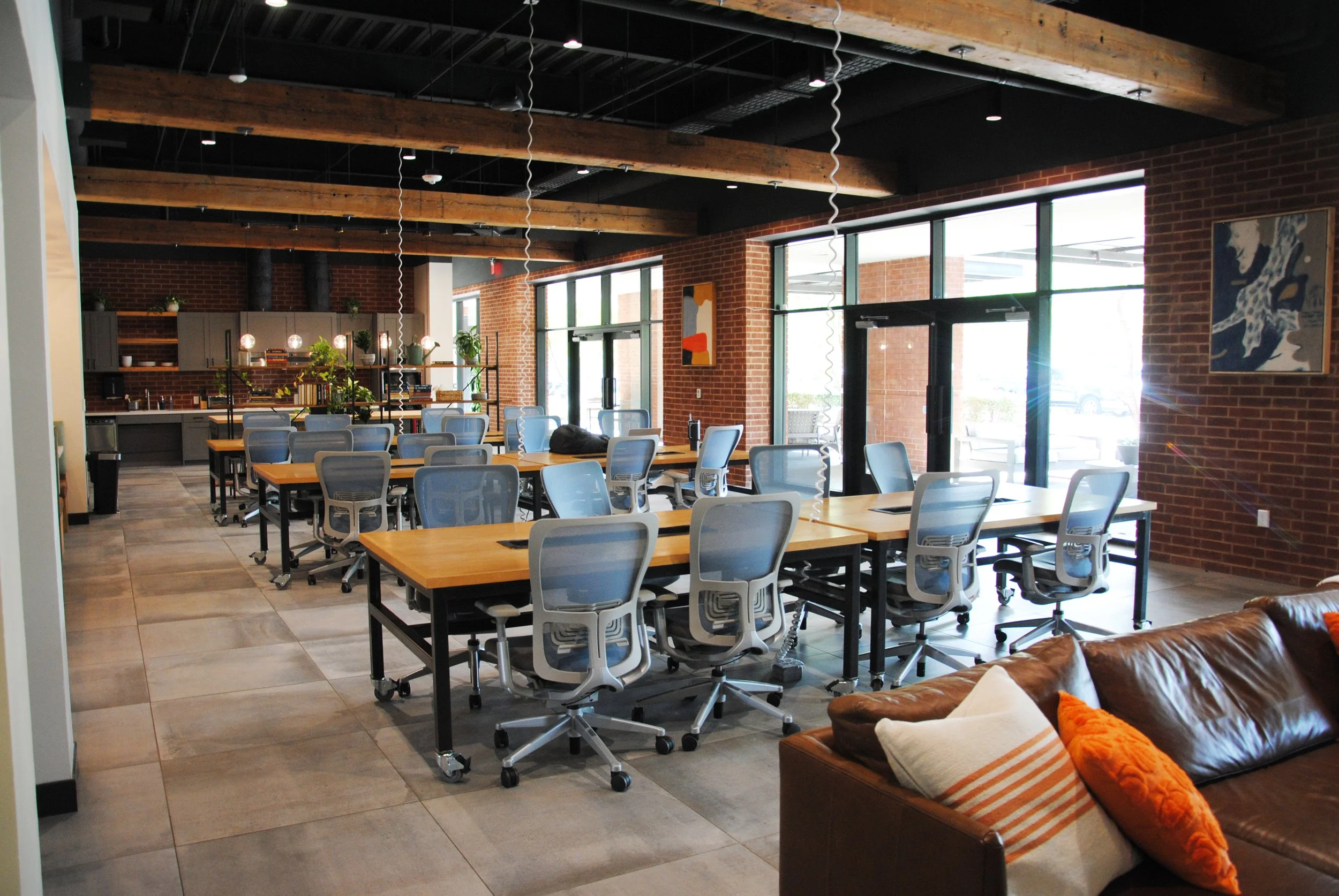 Modern coworking space with multiple light blue ergonomic chairs, wooden tables, a brick wall, large windows, and artwork on the walls.
