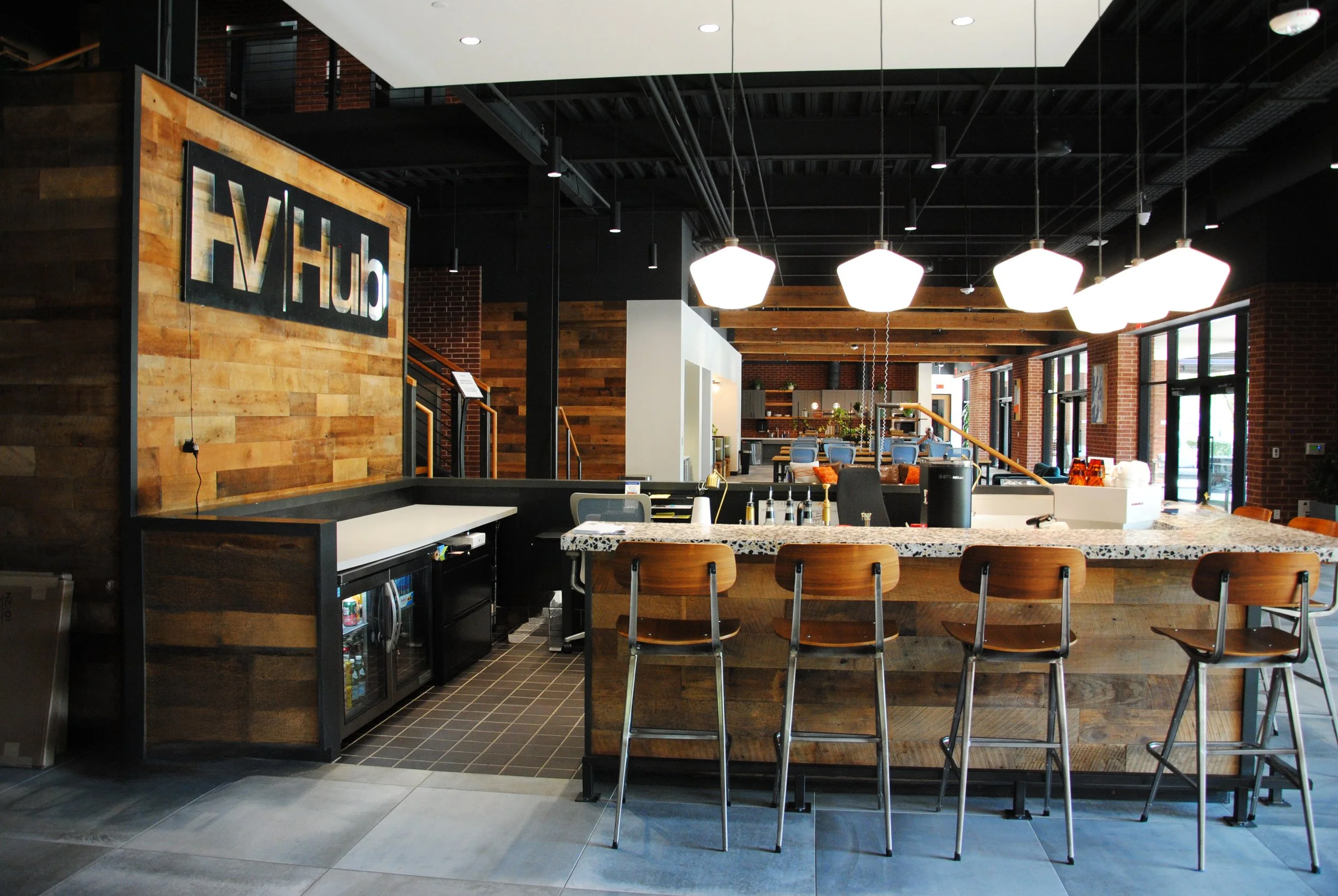 Modern indoor lounge or bar area with a wooden aesthetic, bar stools at a speckled granite counter, pendant lights overhead, and a large sign reading 'Hulu' on a wood-paneled wall.