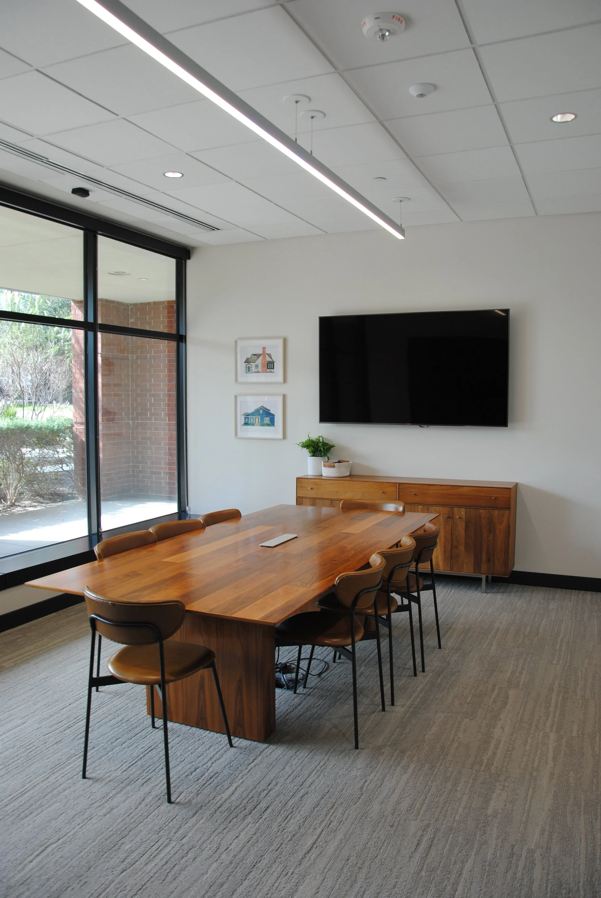Modern conference room with a large wooden table, seven brown chairs, a wall-mounted flat-screen TV, and a wooden cabinet with a plant and decor. Large windows reveal an outdoor brick porch and greenery.