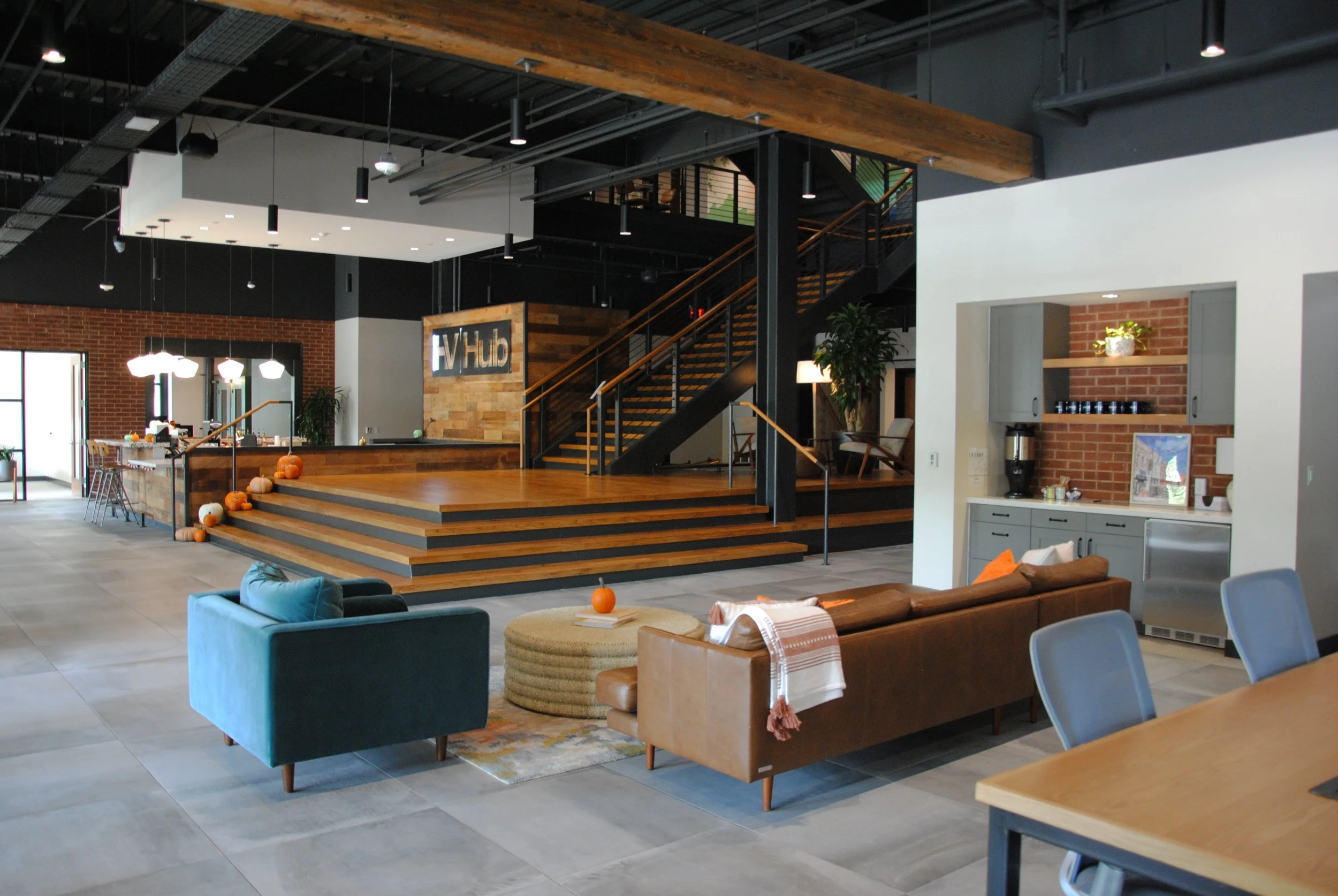 Interior of a modern, open-concept office space with a stage area decorated for fall, seating including a teal armchair and a brown leather sofa, a small round ottoman with a pumpkin, a kitchenette with gray cabinets, and a staircase leading to an upper level.