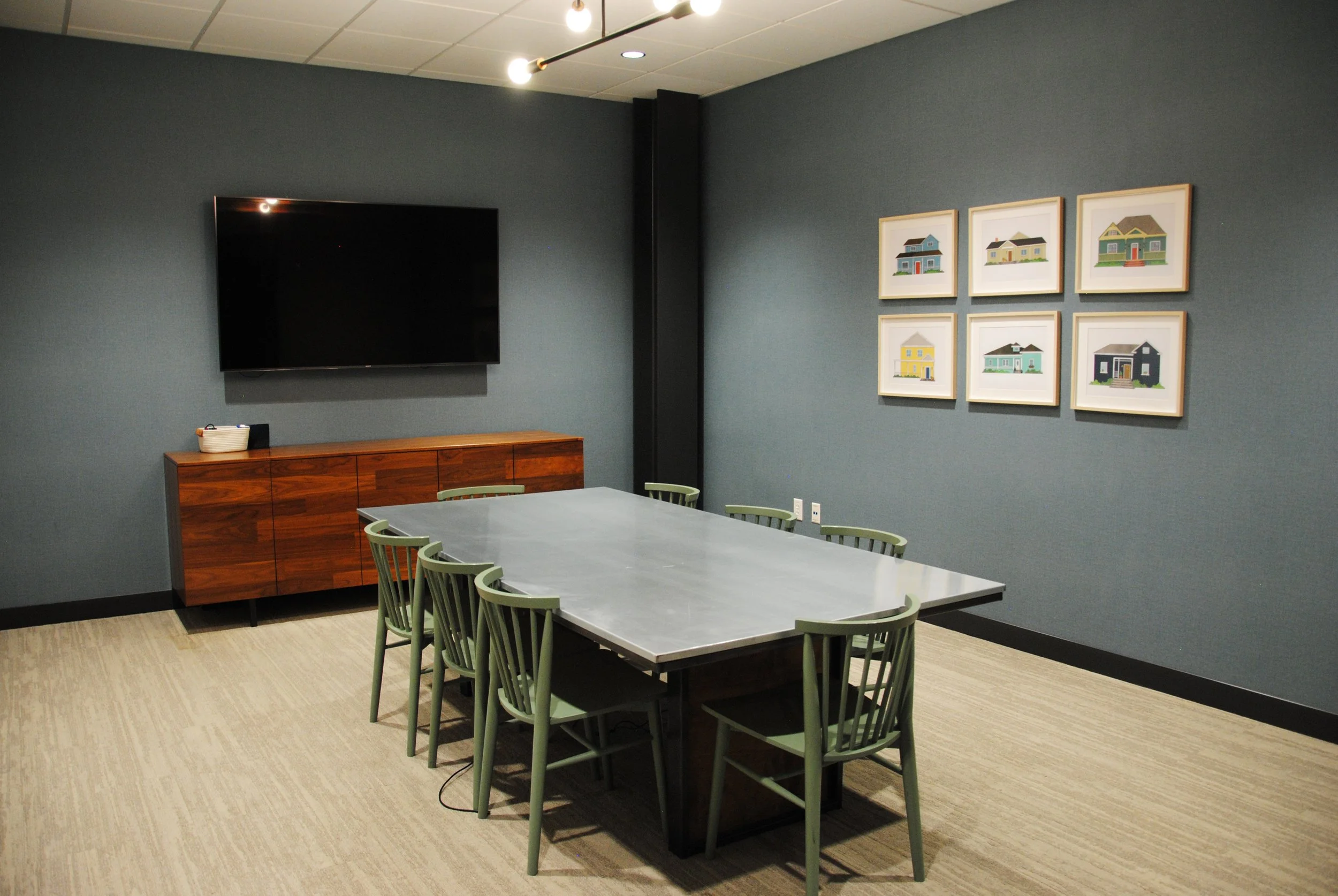 Contemporary conference room featuring a long grey table, green chairs, a dark wood sideboard, a mounted flat-screen television, and six framed colorful house illustrations on a blue wall.