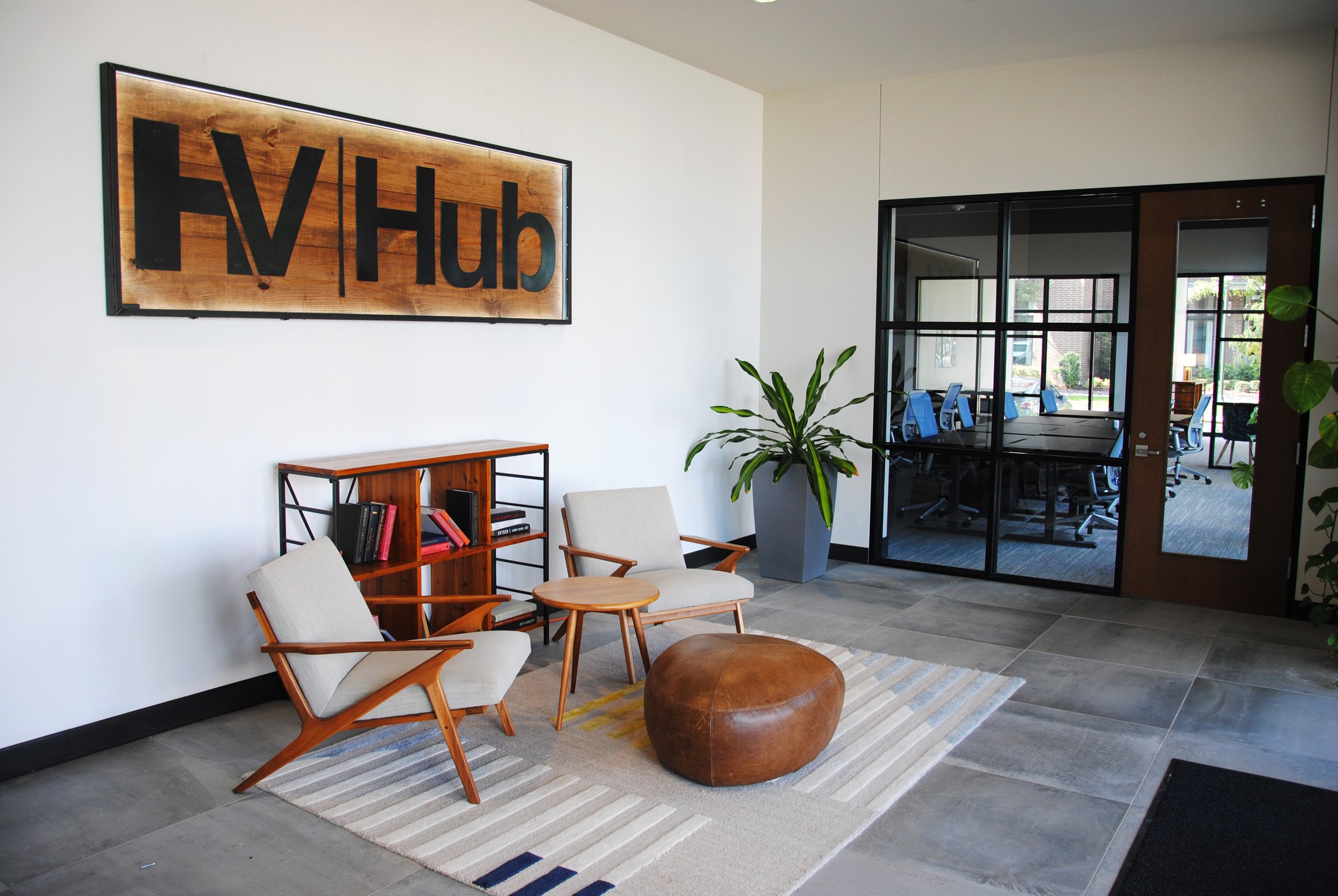 A modern office space with rows of empty desks and ergonomic chairs, large windows letting in natural light, and bookshelf units against the wall with plants and picture frames.