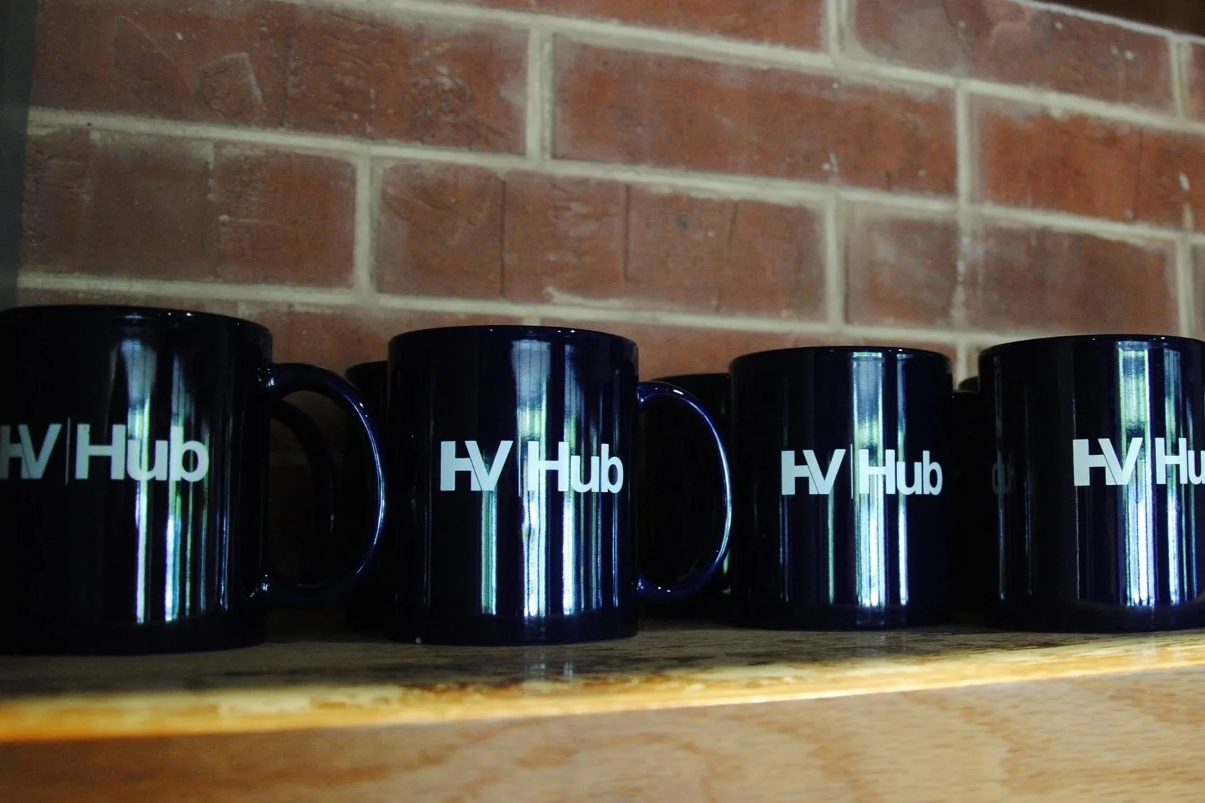 Several black coffee mugs with the logo 'HV Hub' on a wooden shelf against a brick wall.