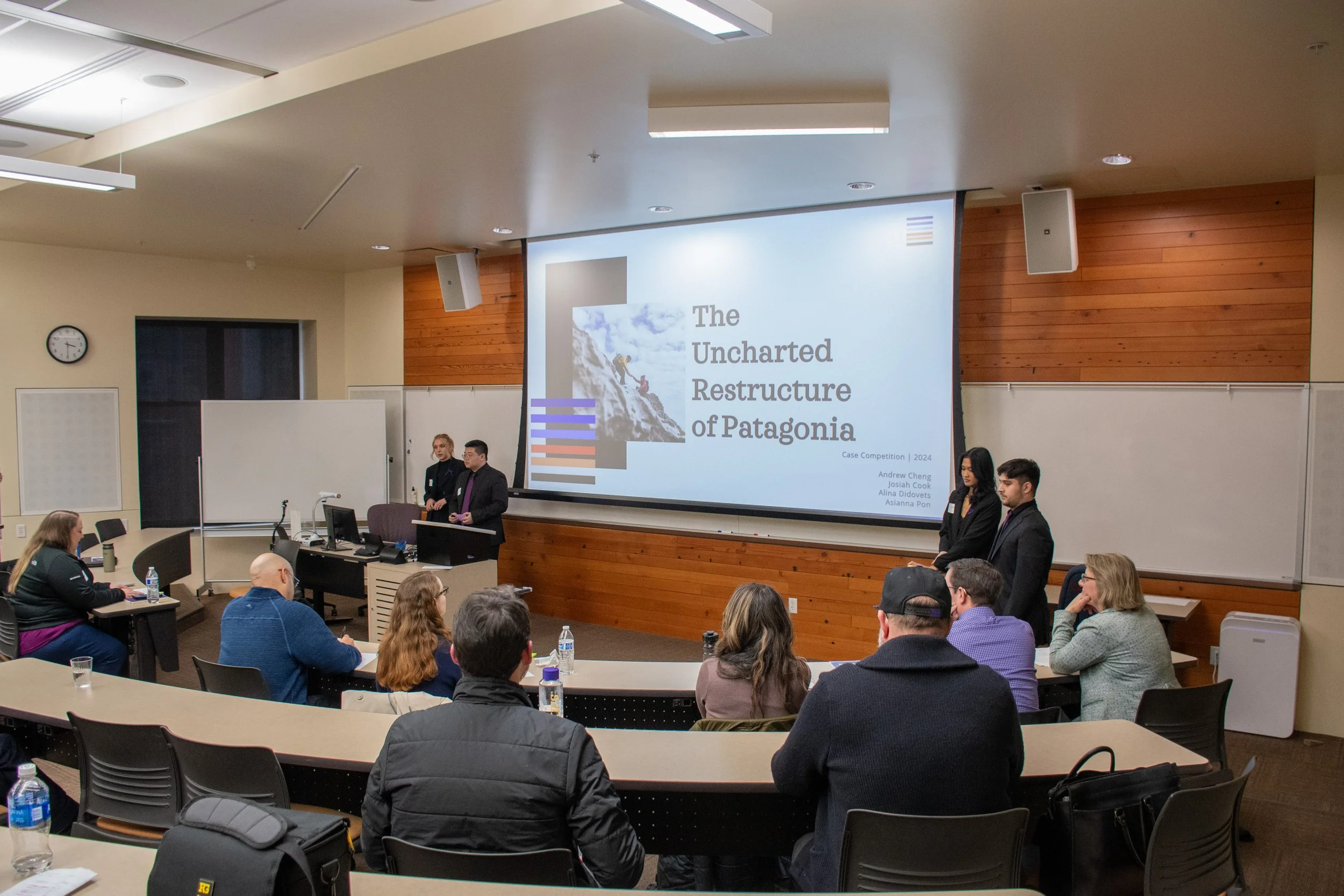 A classroom presentation on Patagonia with four presenters standing in front of a large screen, audience seated at desks, some taking notes, in a university lecture hall.