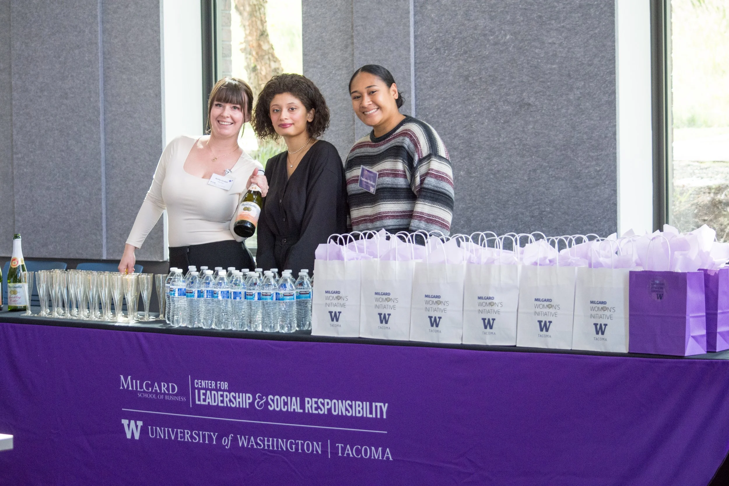 Three women standing behind a table at an event, with water bottles, glasses, and gift bags. The table has a purple tablecloth with the logos of the University of Washington and Milgard School of Business, and the words 'Center for Leadership & Social Responsibility'.