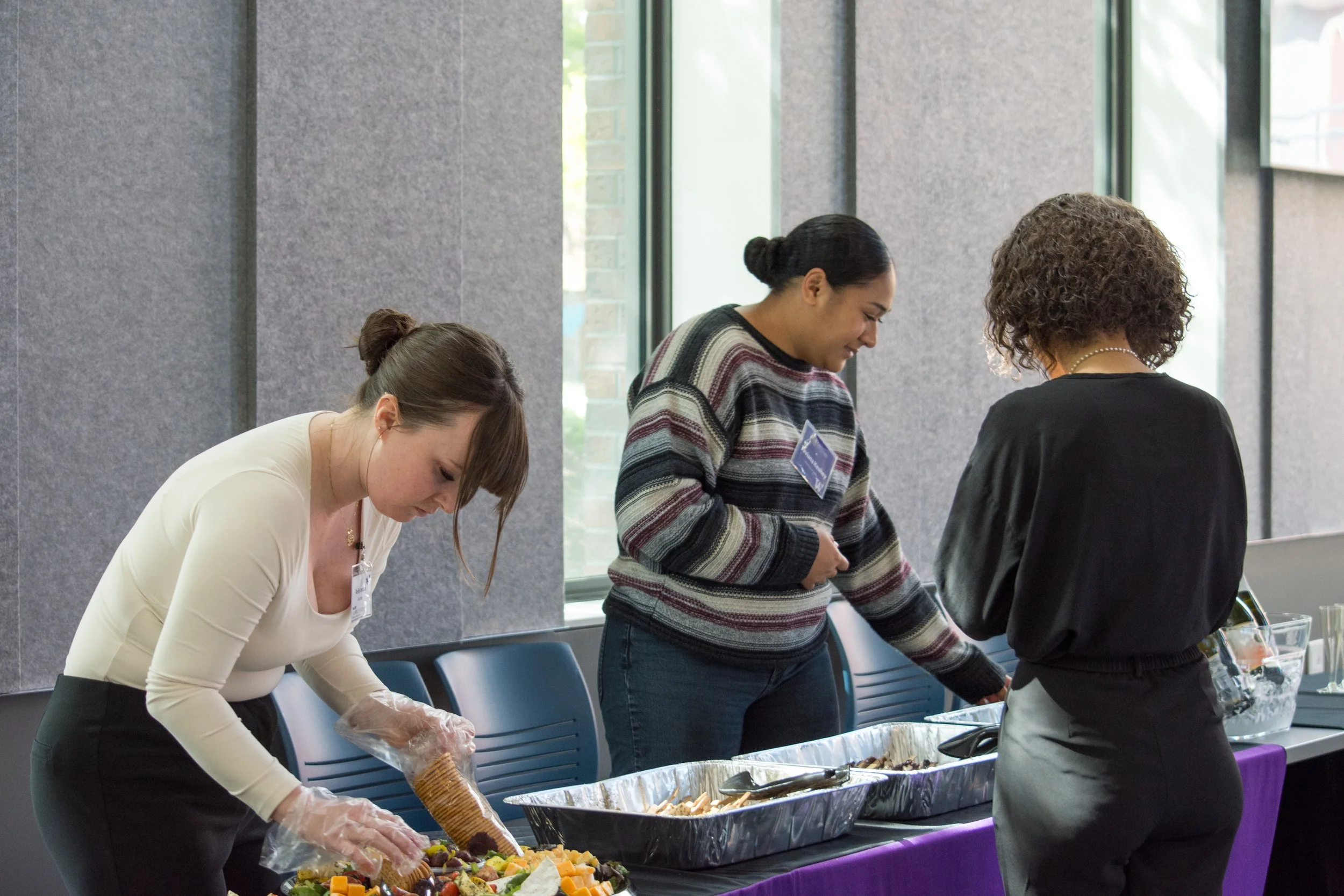 Three women at a buffet table serving themselves food, with food trays and utensils, inside a room with large windows and gray walls.