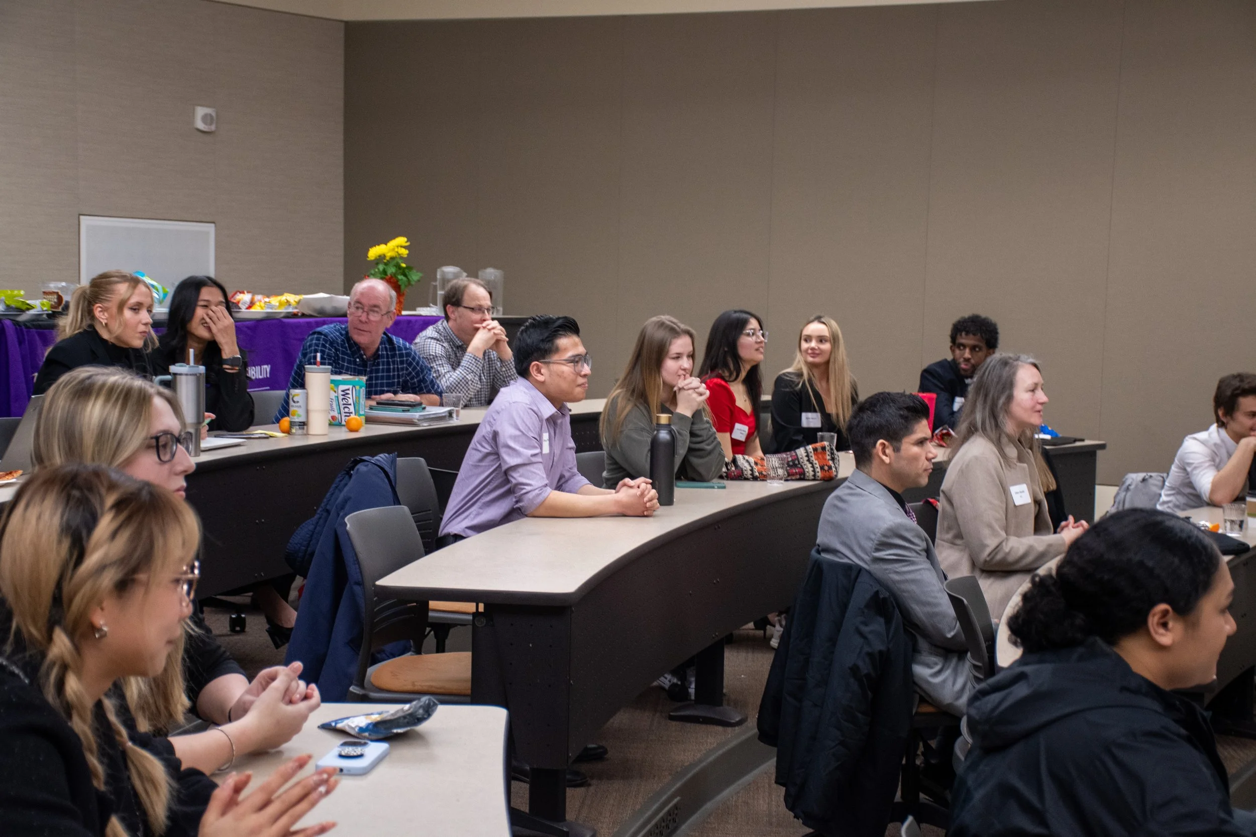 A group of diverse people seated at curved desks in a conference room, listening attentively, with snacks and drinks on a table at the back.