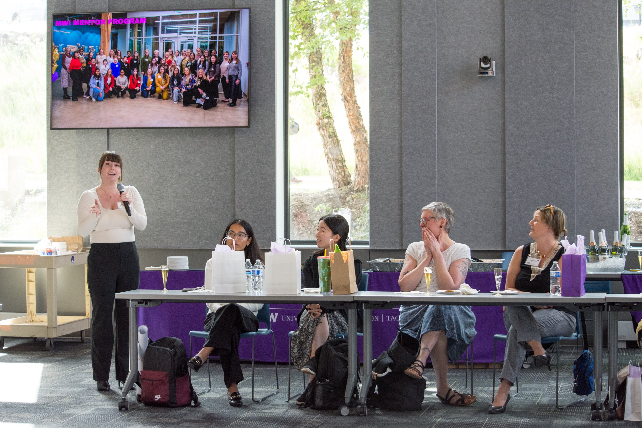 A woman is giving a presentation at a conference or seminar, with four women sitting at a long table listening. A large screen displays a group photo with the title 'MWI Mentor Program'.
