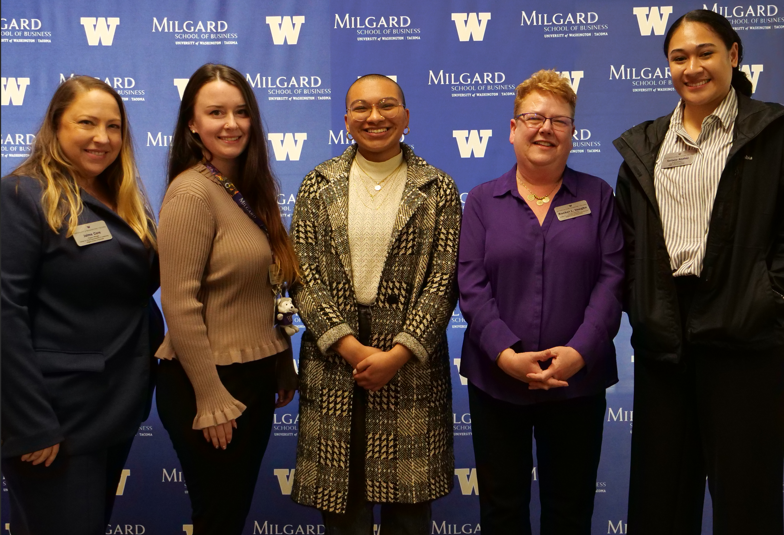 Five women standing together in front of a blue backdrop with 'Milgard School of Business' and the University of Washington logos, smiling for a group photo.