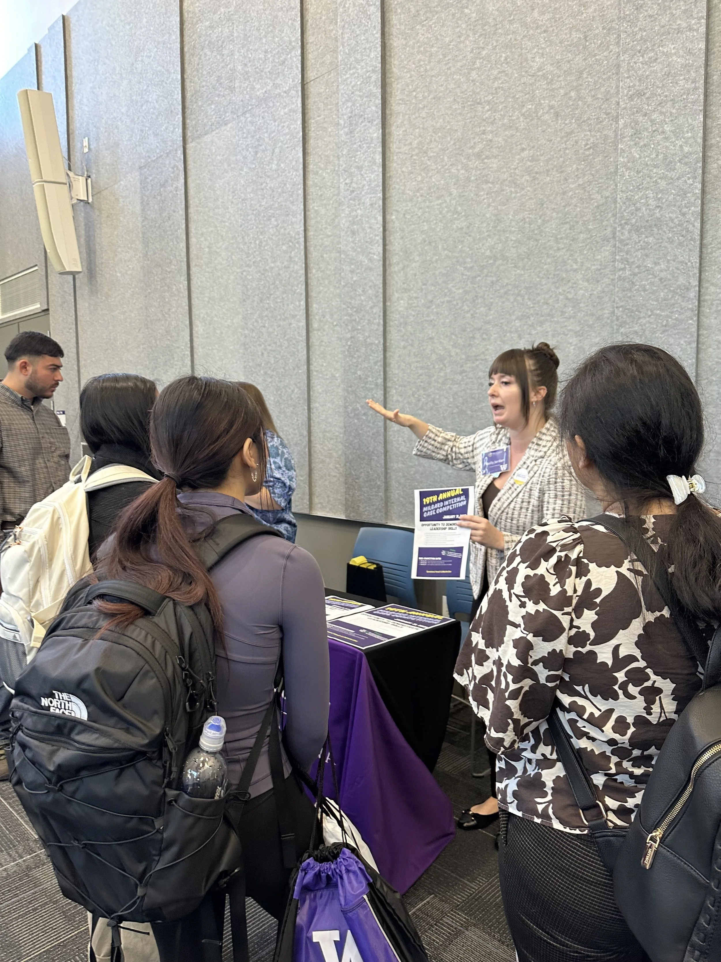 A woman is speaking to a group of people at an indoor event, possibly a conference or a fair, with informational materials on the table in front of her.