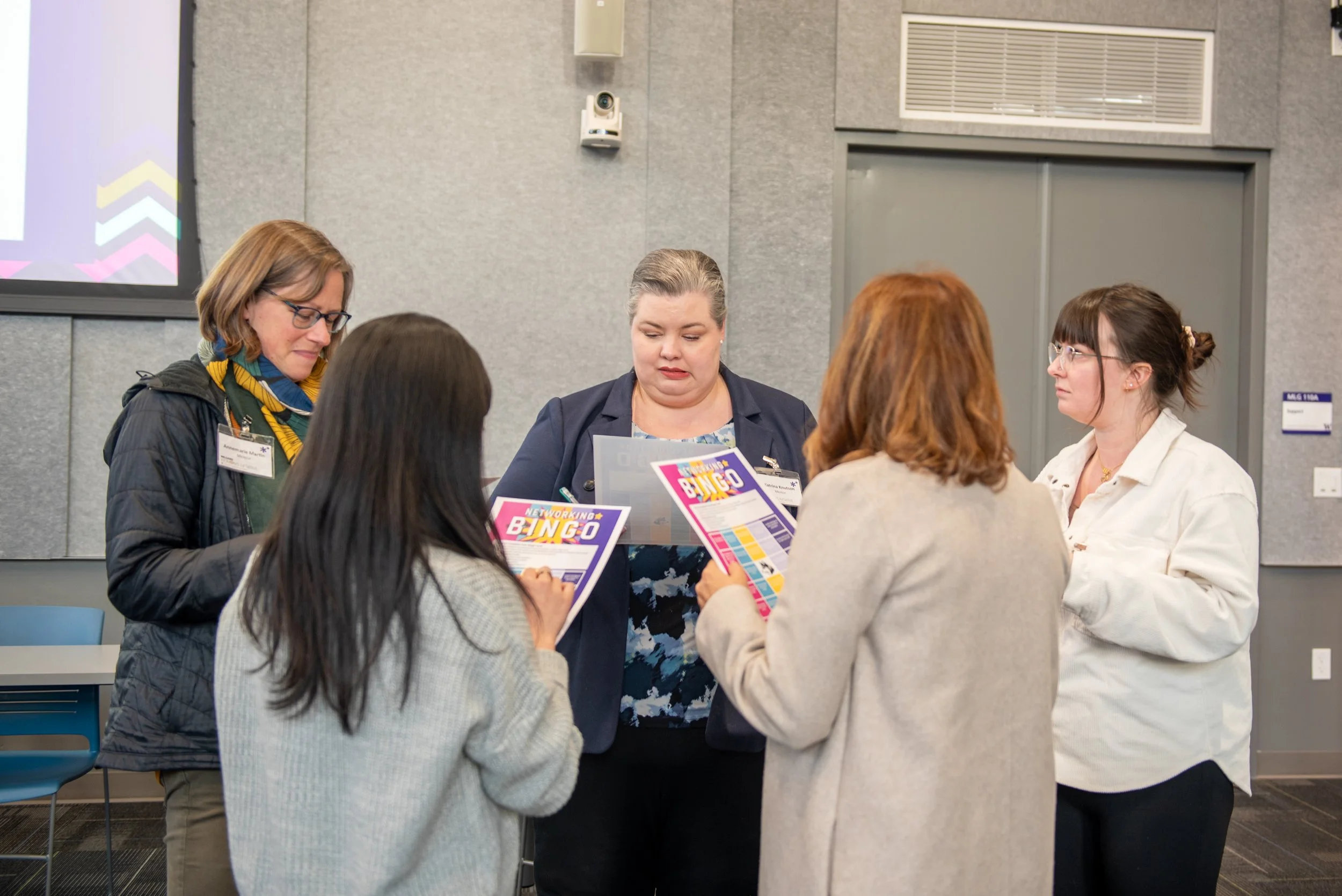 Five women in a group discussion at an indoor conference, some holding colorful bingo papers, with gray walls and a projector screen in the background.