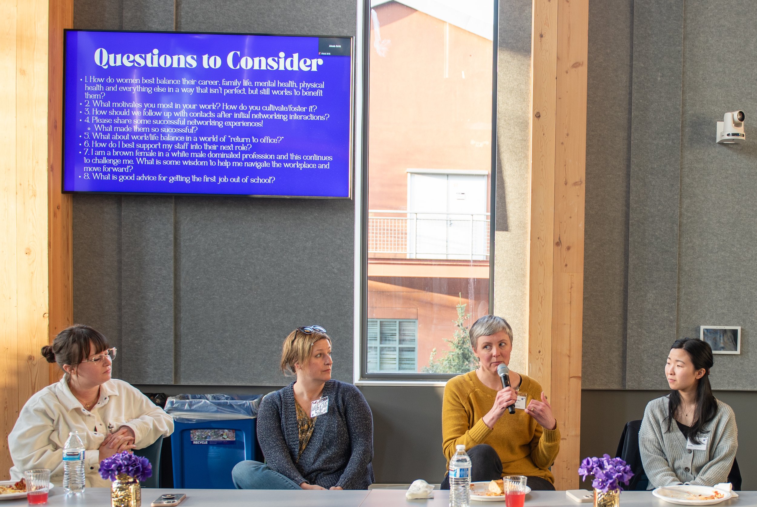 Four women sitting at a table during a panel discussion in a room with wood and gray walls. One woman is speaking into a microphone, and a large screen behind them displays questions to consider.