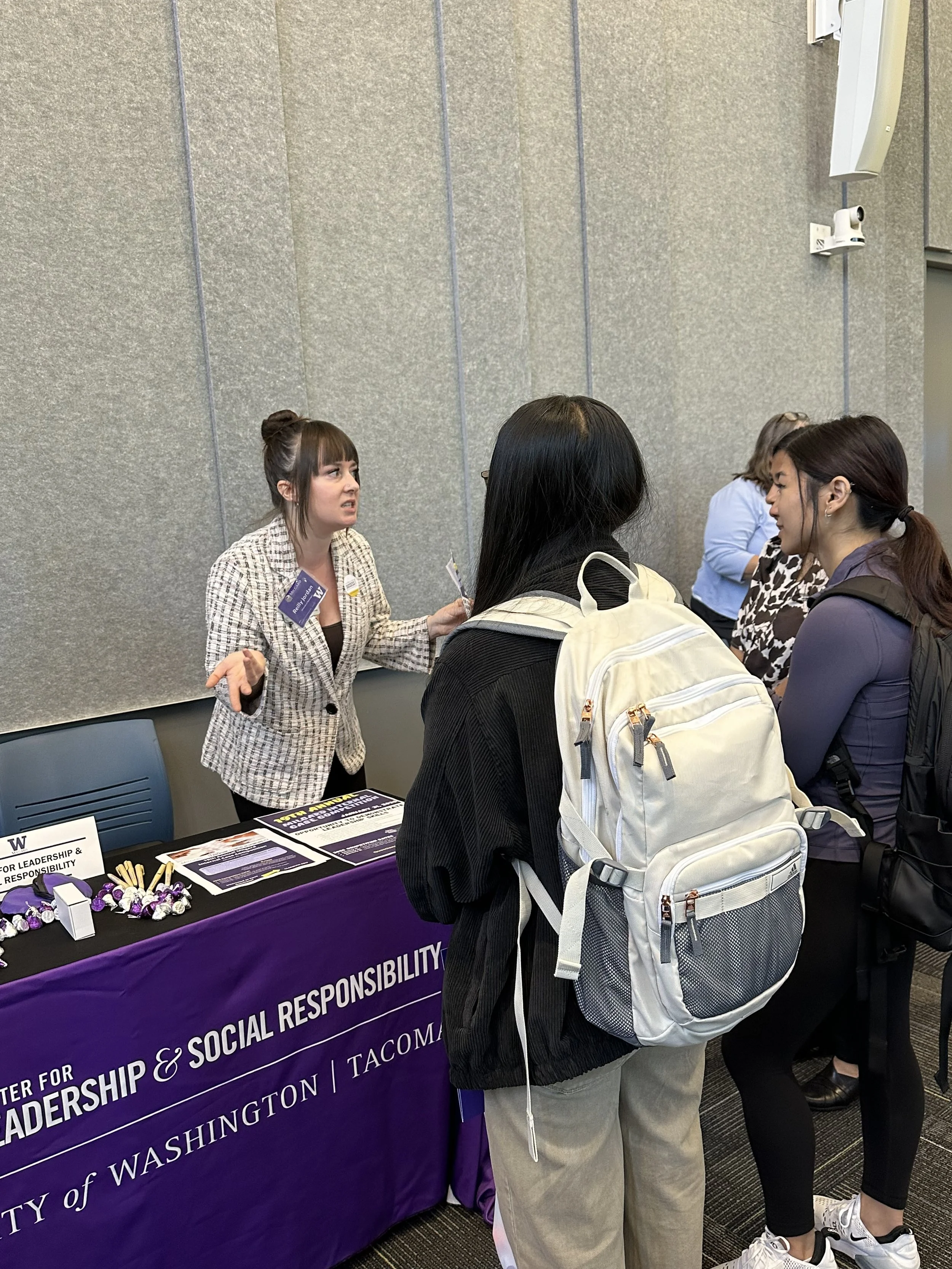 A woman at a conference booth talking to two attendees, with brochures and purple branded merchandise on the table, representing the University of Washington Center for Leadership & Social Responsibility.