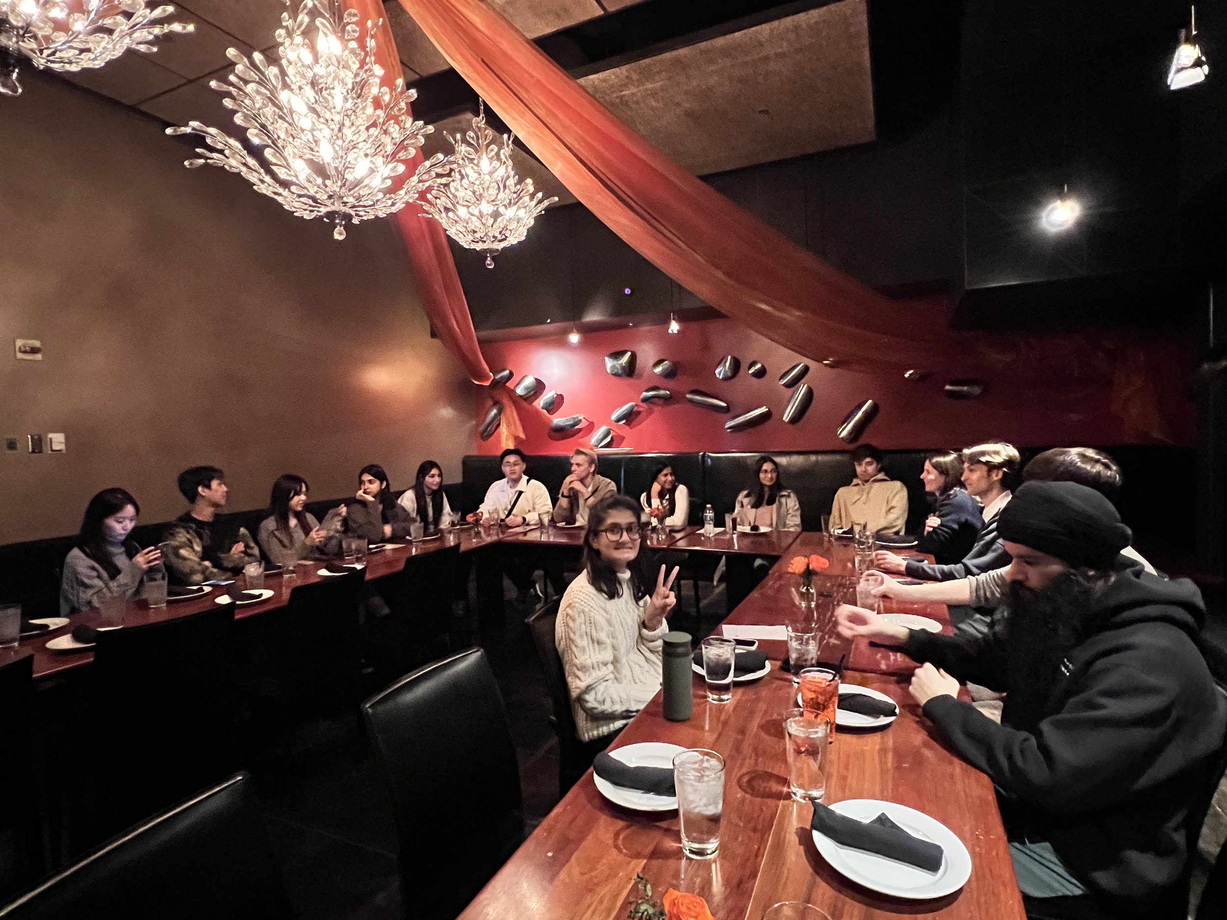 Group of people sitting at a long banquet table in a dimly lit restaurant, with decorative lighting and red fabric drapes on the wall.