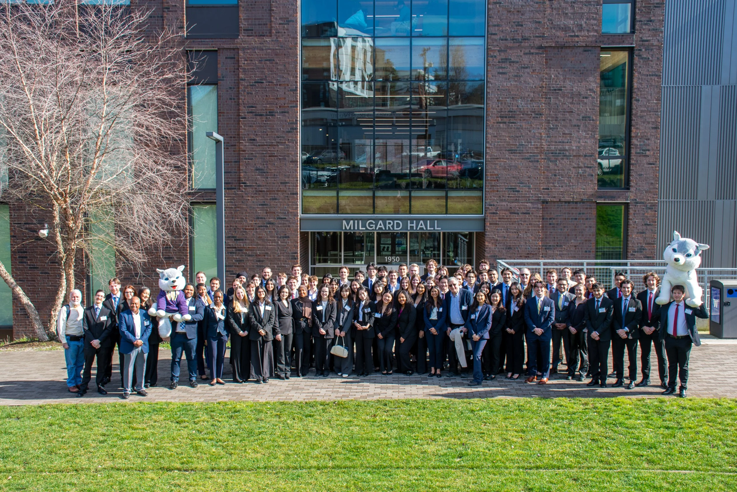 A large group of people, some in suits, standing in front of Milgard Hall at a university or similar institution. Two individuals are wearing large cartoon-like mascot costumes resembling a white cat or dog.