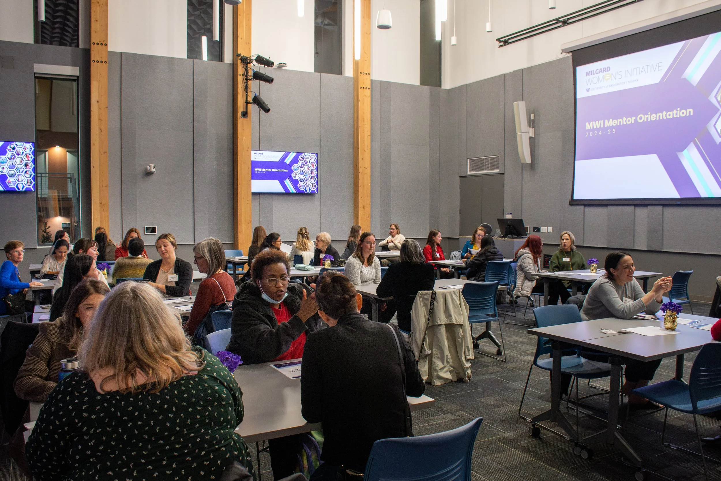 Women sitting at tables in a conference room during an event titled 'MWI Mentor Orientation'. Large screens display the event title, and participants are engaged in conversation.