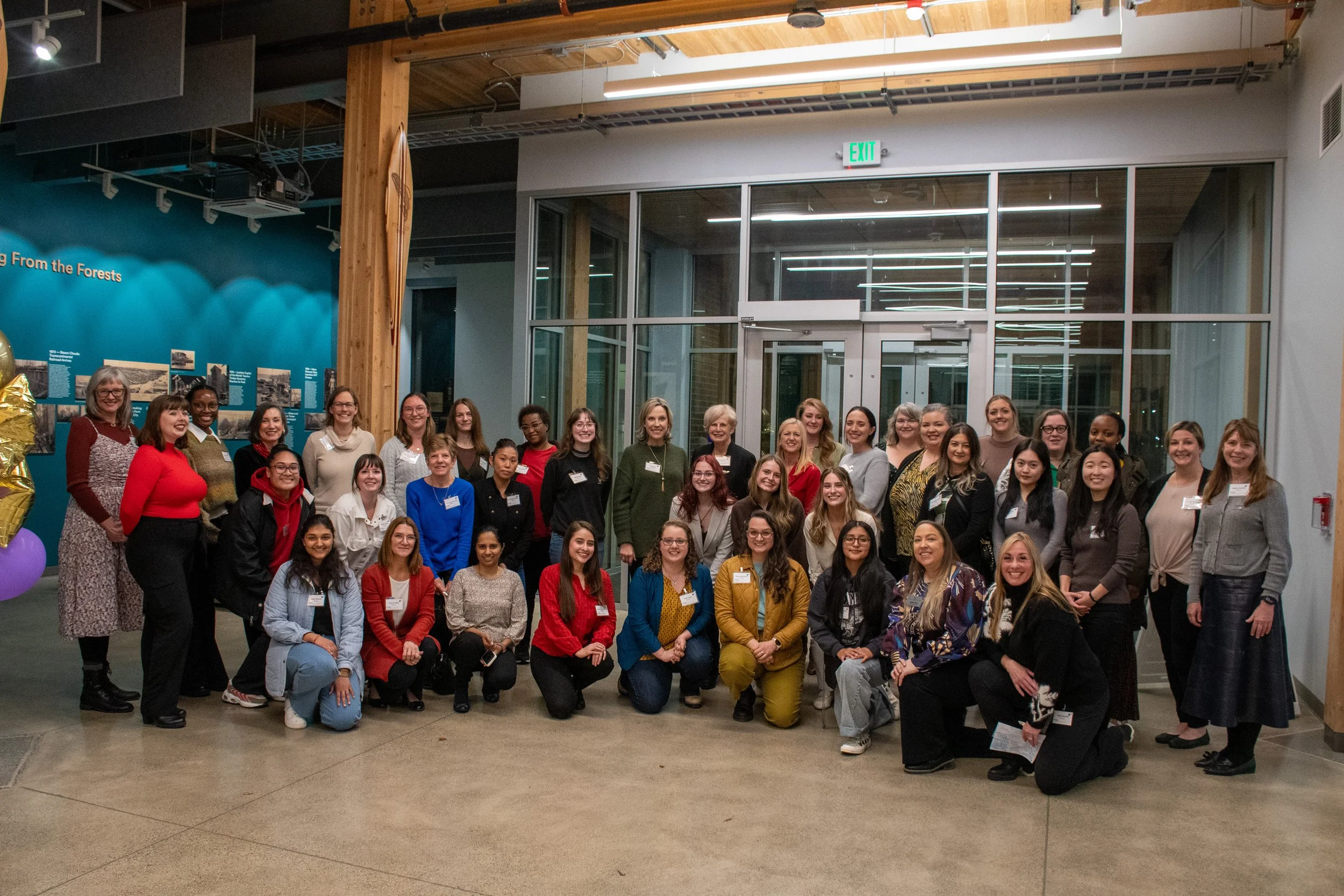 Group of diverse women and girls gathered indoors for a photo, standing on a concrete floor in front of a glass entrance and a blue exhibit wall with informational plaques.