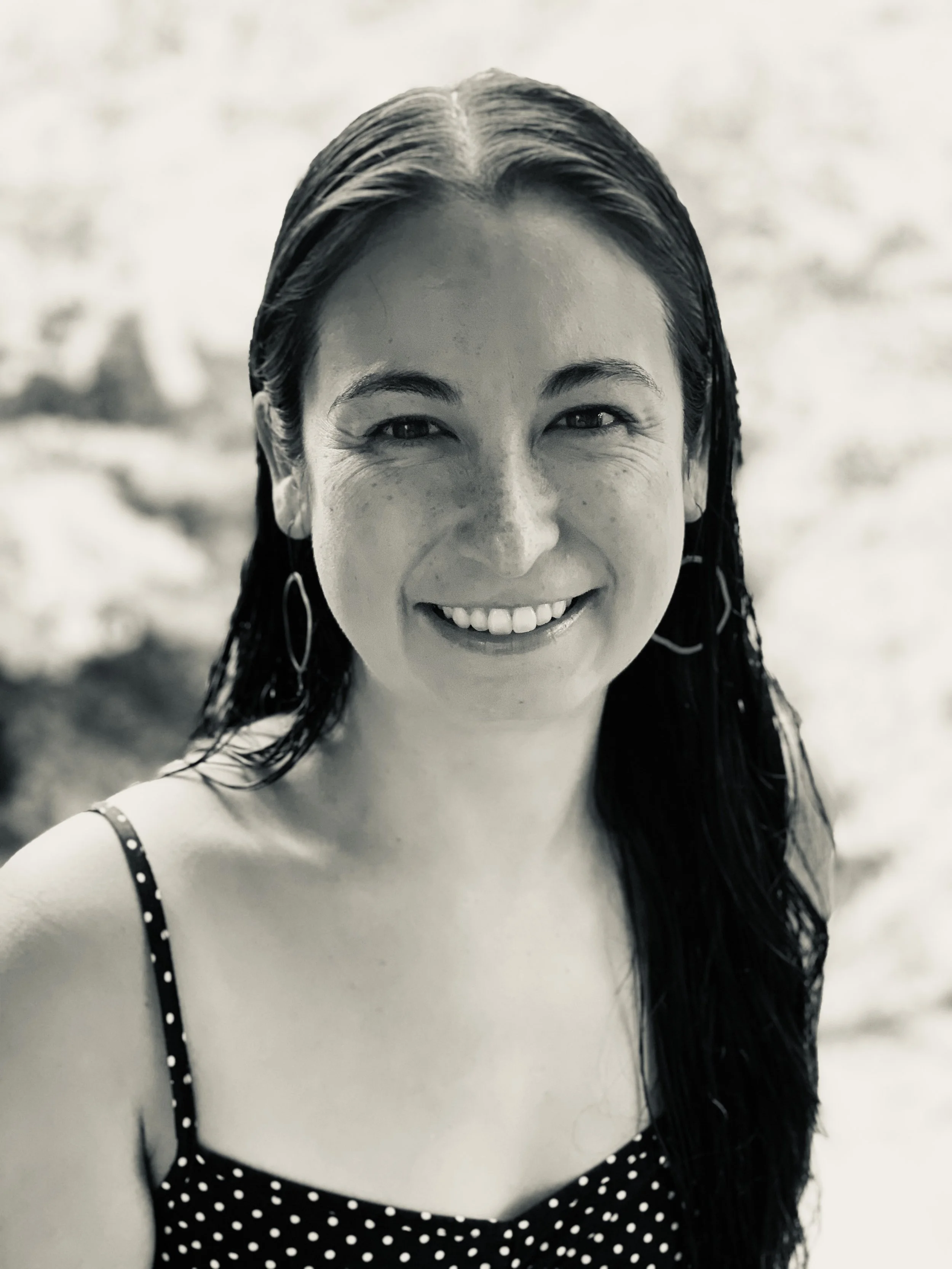 Black and white photo of a smiling woman with wet hair, wearing a polka dot swimsuit or top with shoulder straps, standing outdoors.